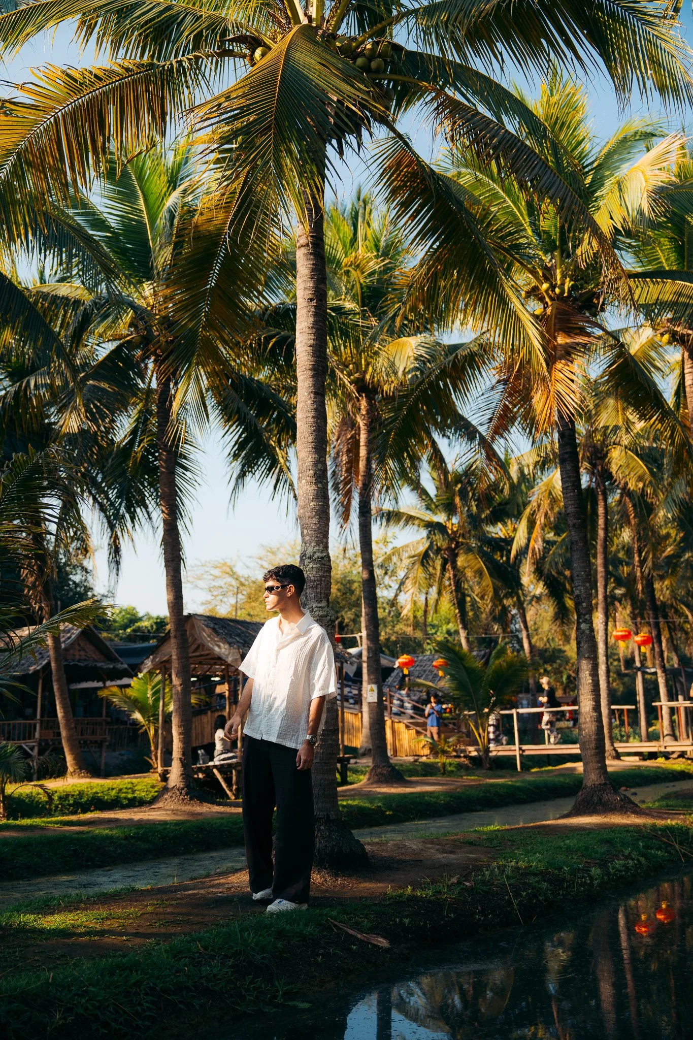 Un hombre con gafas y camisa blanca de manga corta está de pie frente a palmeras en un entorno tropical, con casas de madera tradicionales y faroles rojos decorativos en el fondo, durante el día.