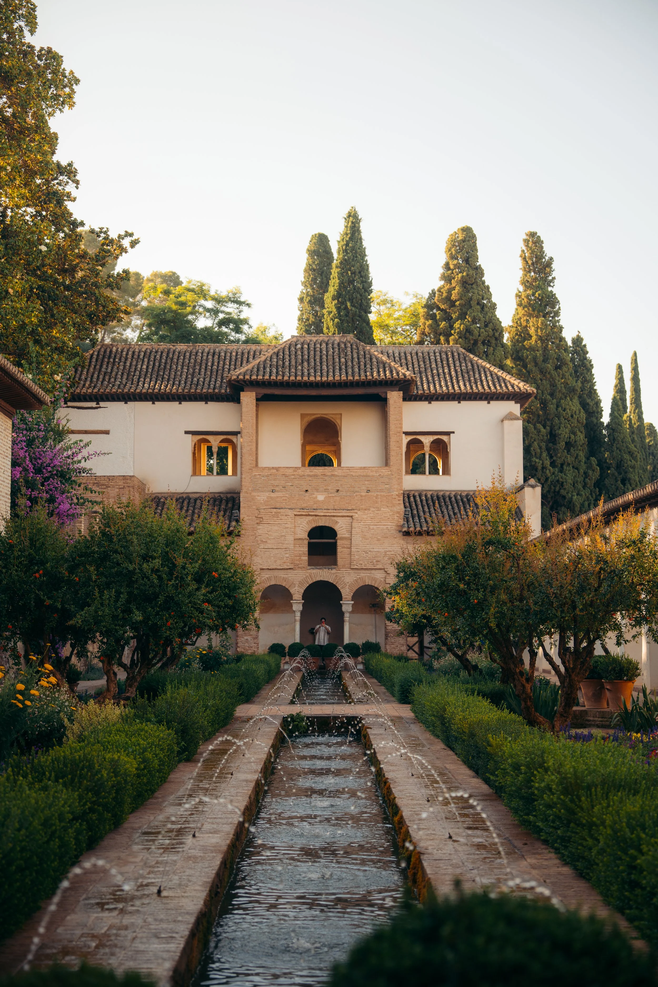 Un edificio con paredes blancas, techos de tejas y arcos en la entrada, rodeado de árboles, un jardín bien cuidado y una fuente central en un estilo arquitectónico tradicional.