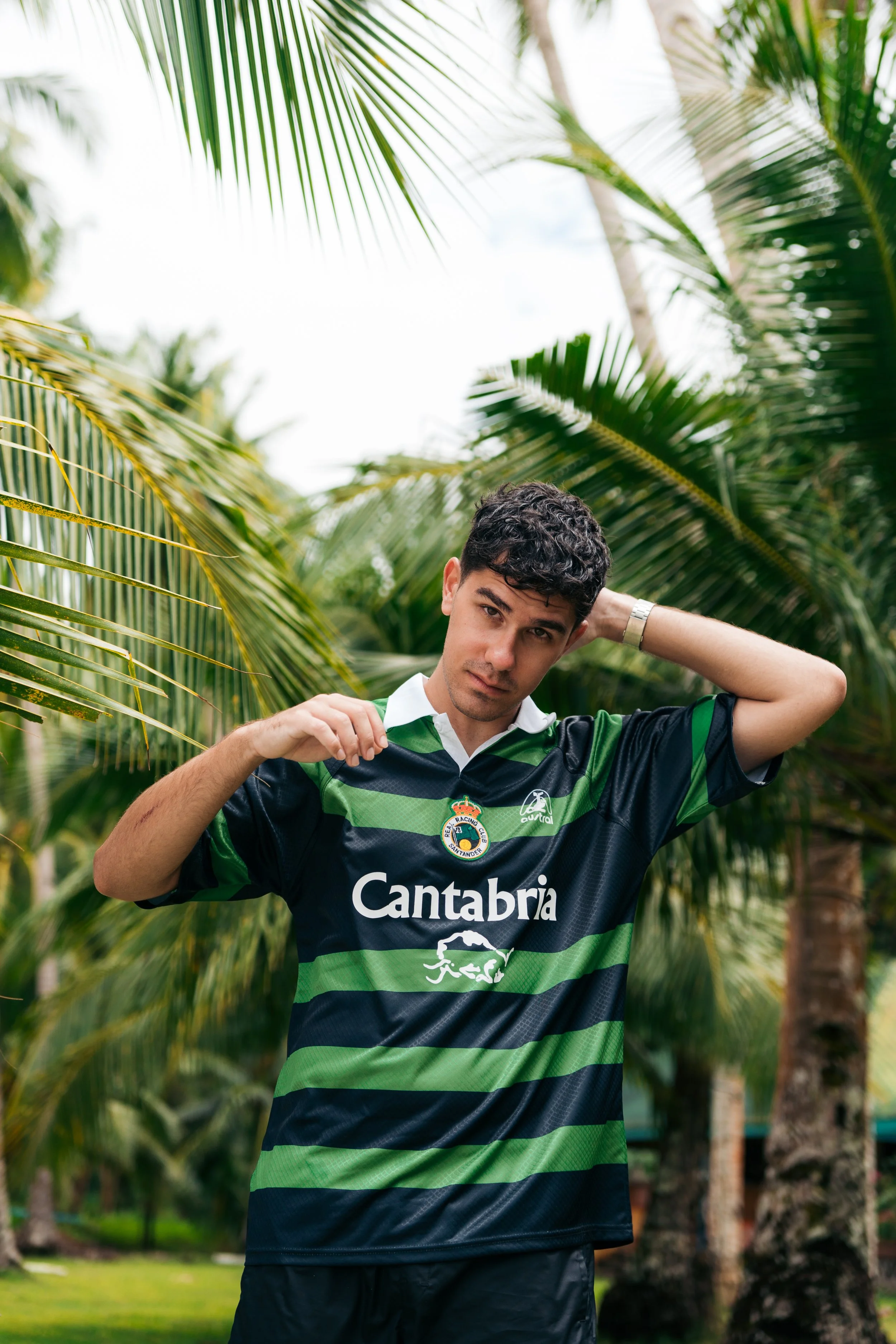 Joven con camiseta de fútbol de rayas verdes y negras, con el logo de Cantabria, posando entre palmeras.