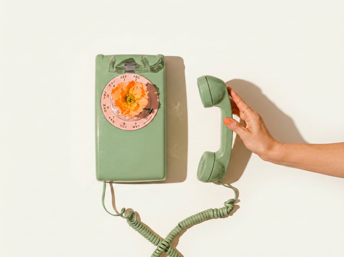 A vintage green rotary telephone with a flower in the center of the dial pad, with a hand holding the receiver on a white background.