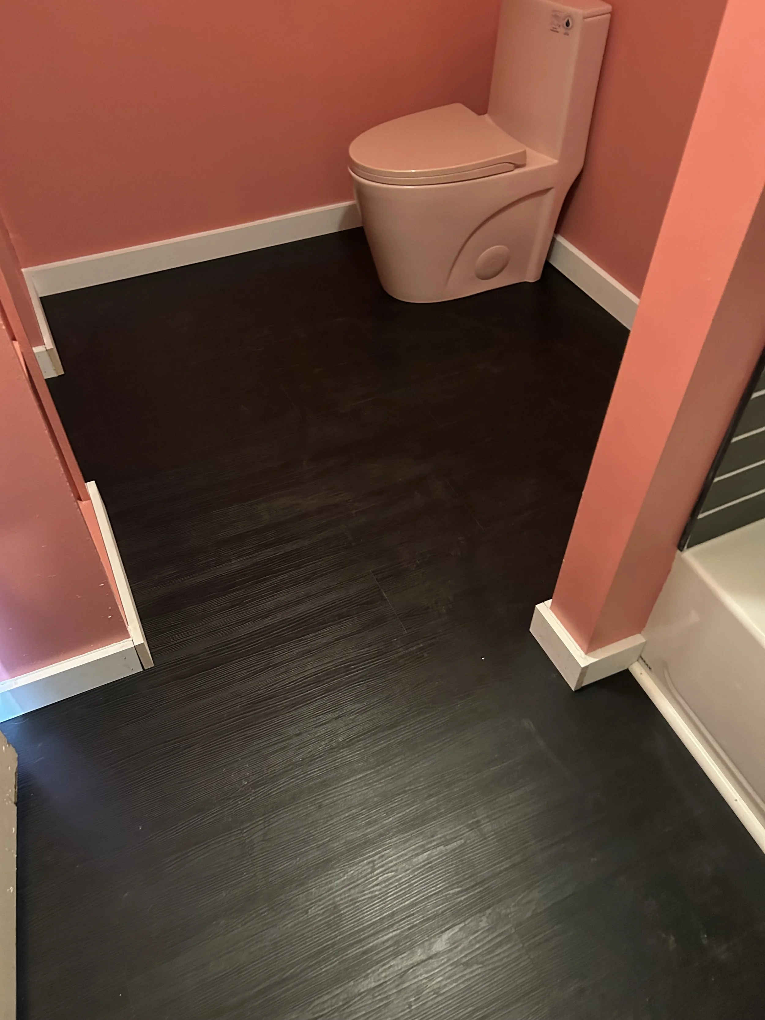 A corner of a bathroom with a white toilet against pink walls, dark wood flooring, and white baseboards.