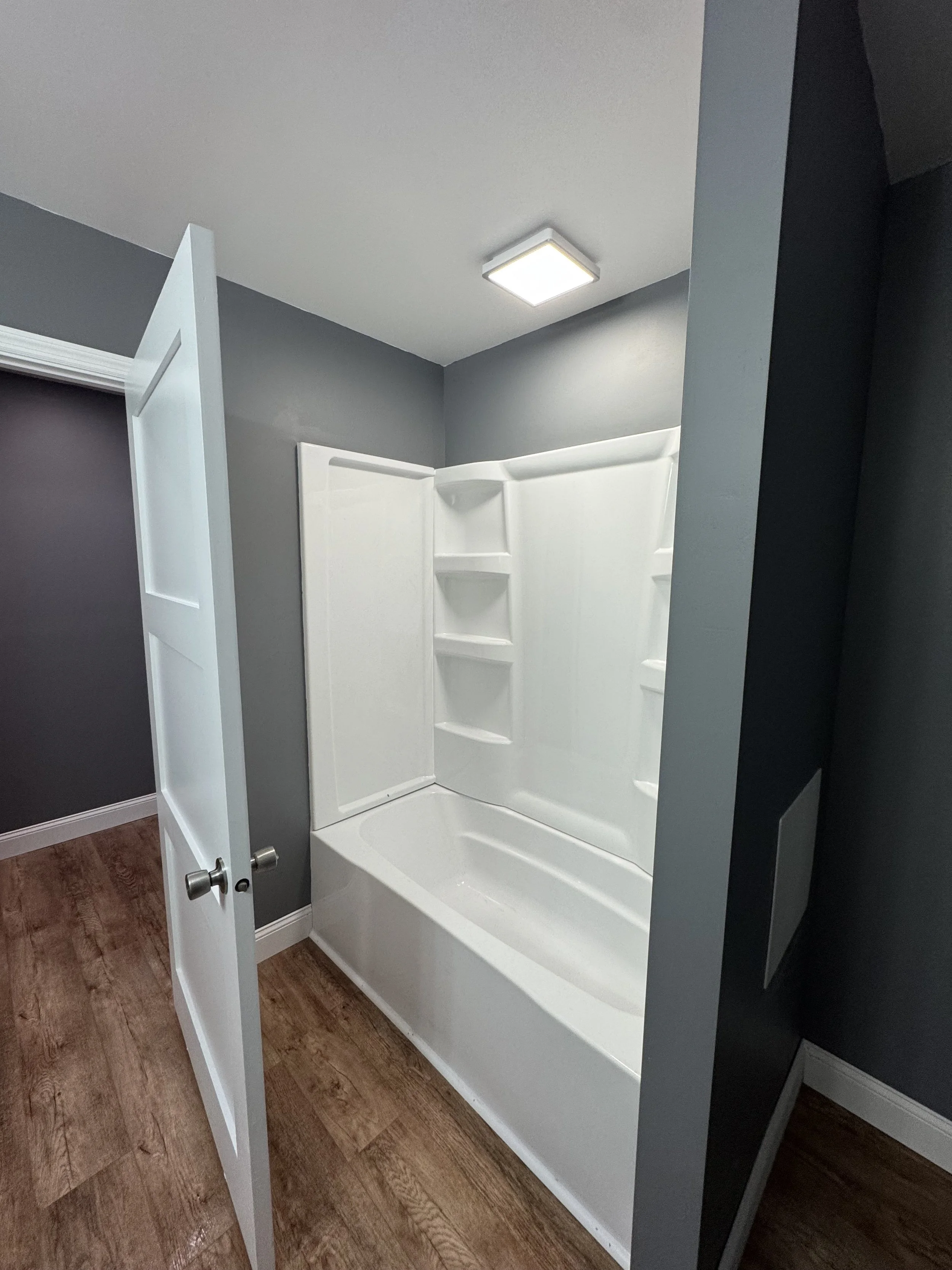 Bathroom shower with white enclosure, opened door, gray walls, ceiling light, wooden floor.