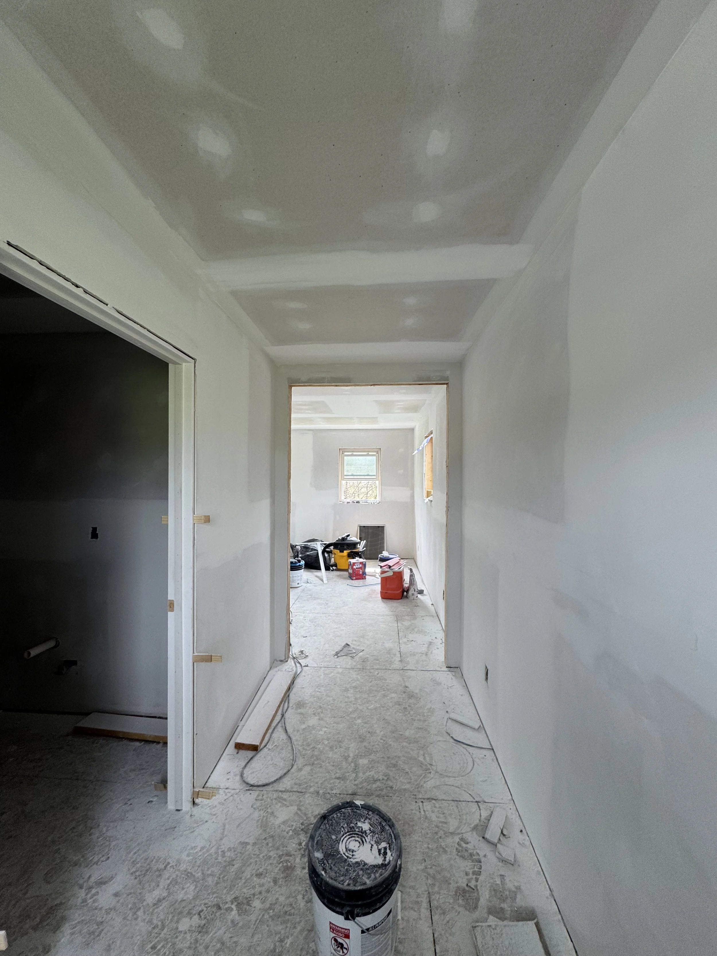 A hallway under construction with drywall installed on the walls and ceiling, tools and supplies scattered on the floor, including a bucket and cans of paint or primer, and an open doorway leading to another room with a window.