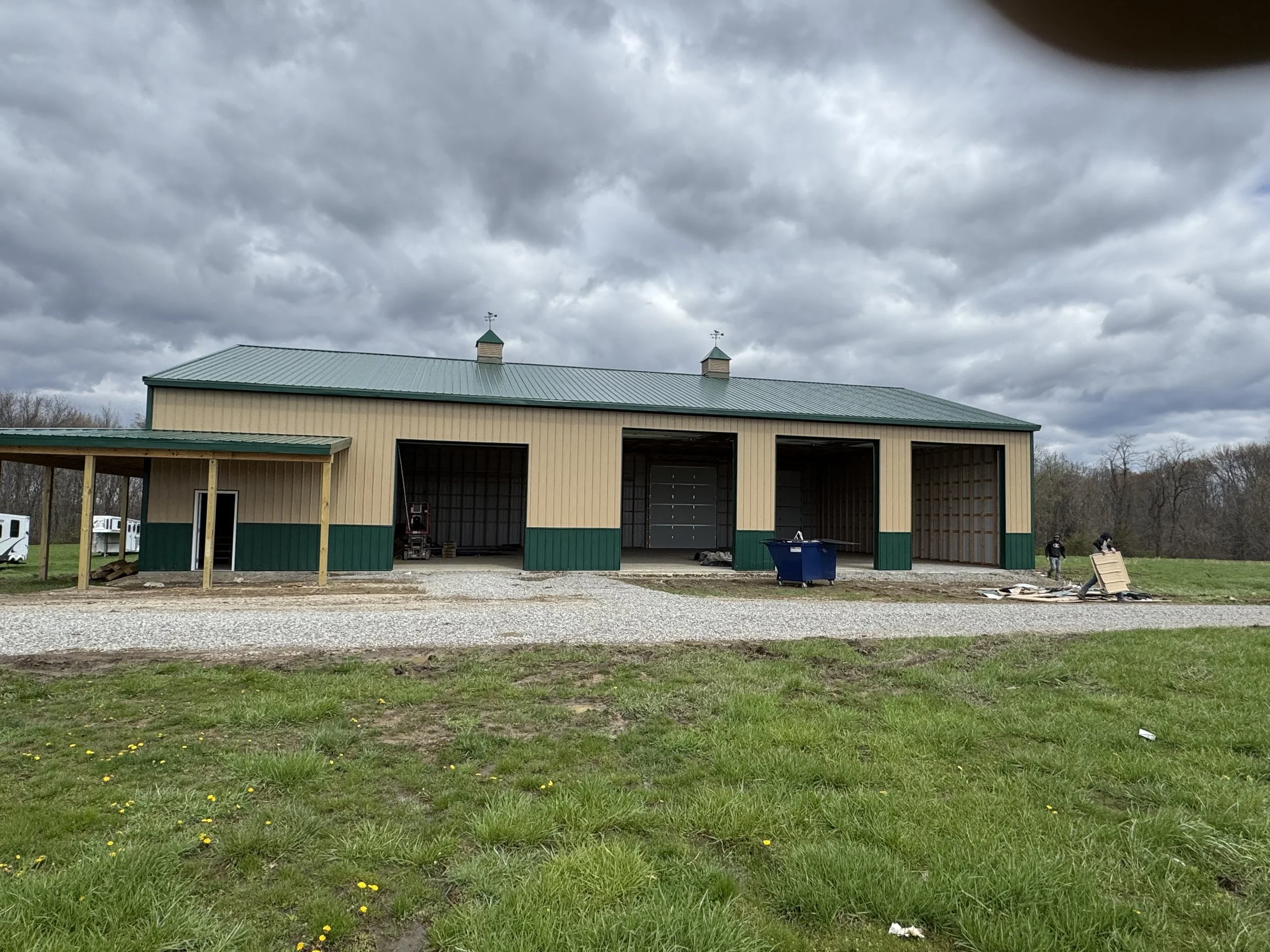 A green and beige steel building under construction, with open garage doors, a grassy area in front, and cloudy skies overhead.