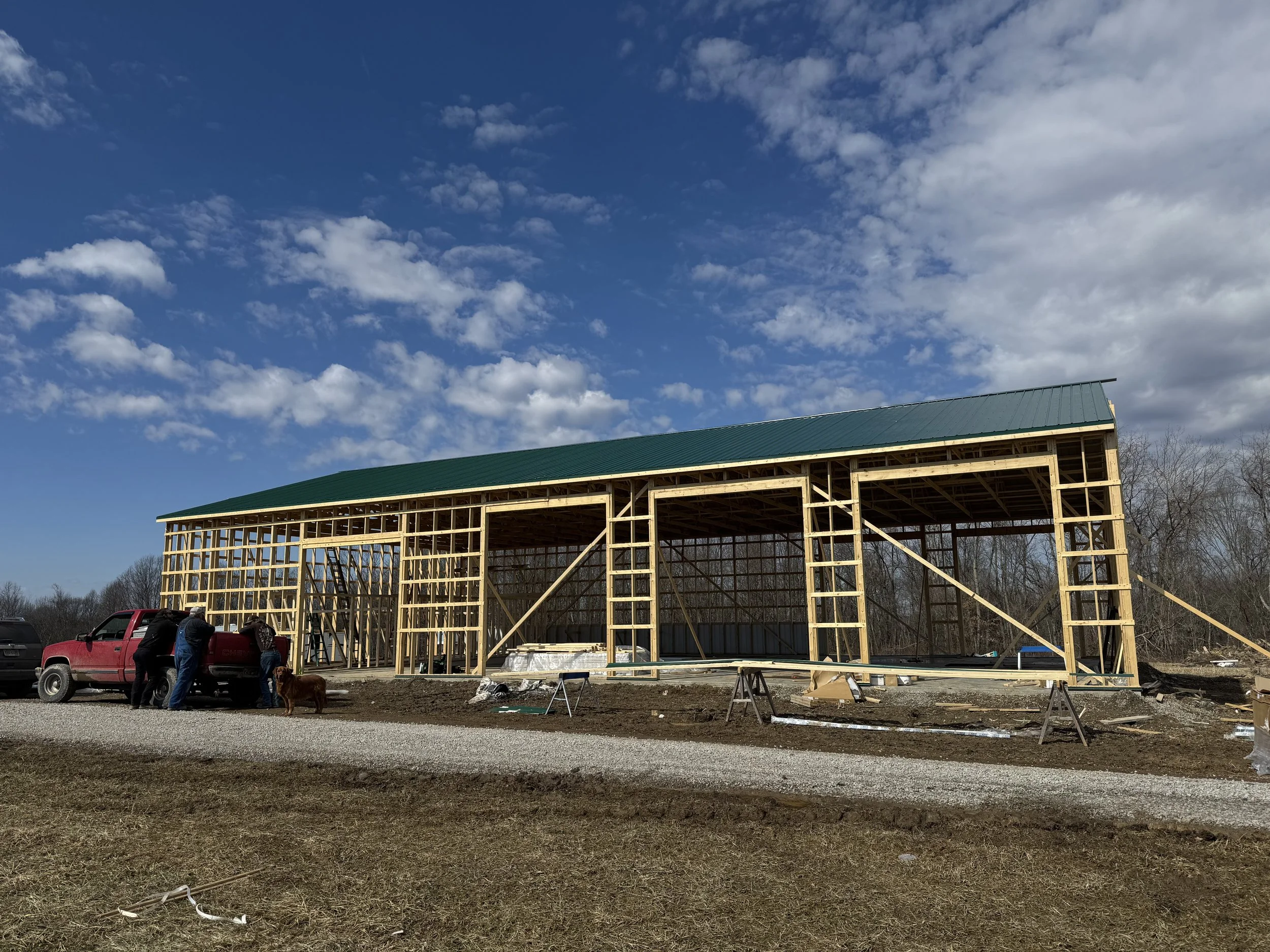 A partially constructed wooden barn with a green metal roof under a blue sky with scattered clouds. There are workers and a red pickup truck at the building site, with construction materials and tools scattered around.