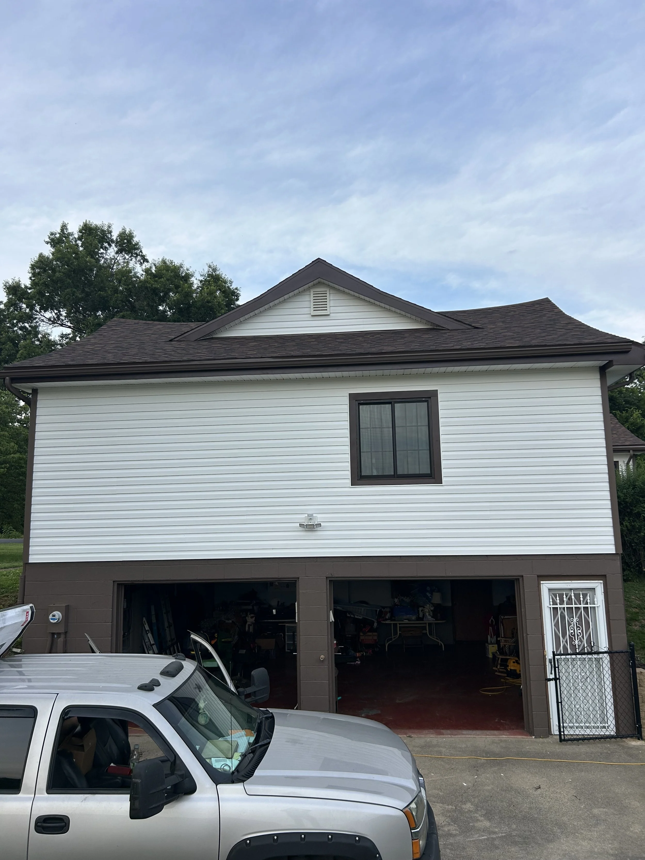 A two-story house with white siding on the upper portion and brown on the lower. It has a gabled roof and a window on the upper floor. The house has a garage with two open bays filled with tools and equipment. A silver vehicle is parked in front of the house, and a fence is visible on the right side.