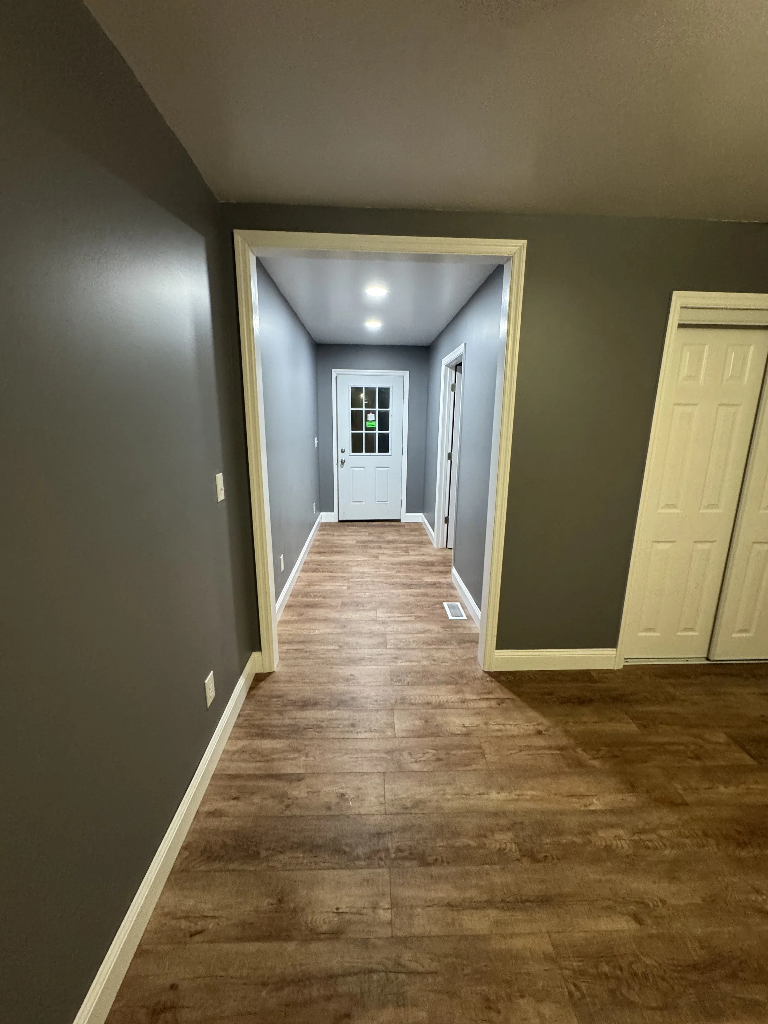 Interior view of a hallway with wood flooring, gray walls, white trim, and a white door at the end of the hall.