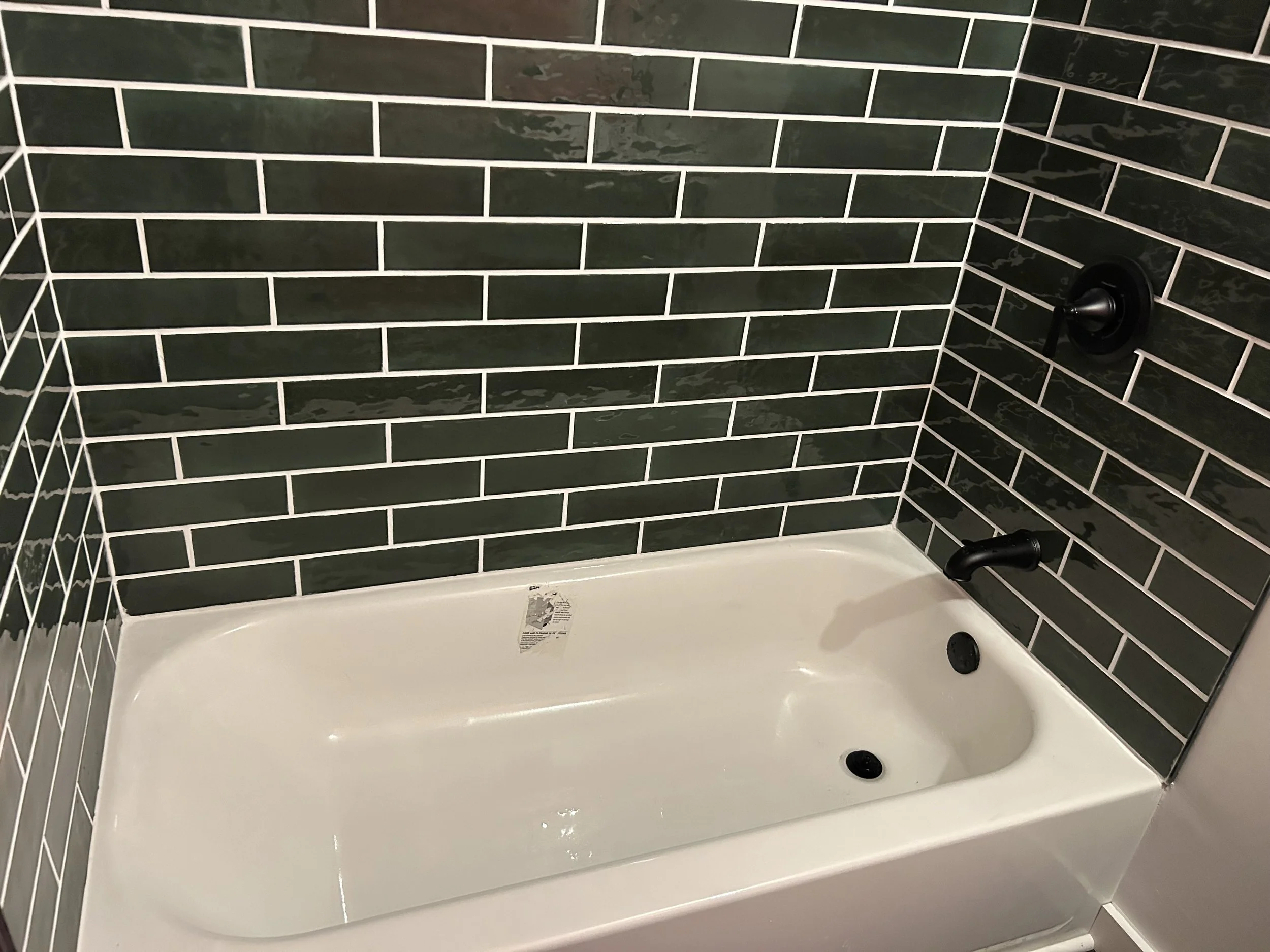 A white bathtub with black fixtures, surrounded by dark green subway tiles on the walls, and a black drain stopper in the bottom right corner.