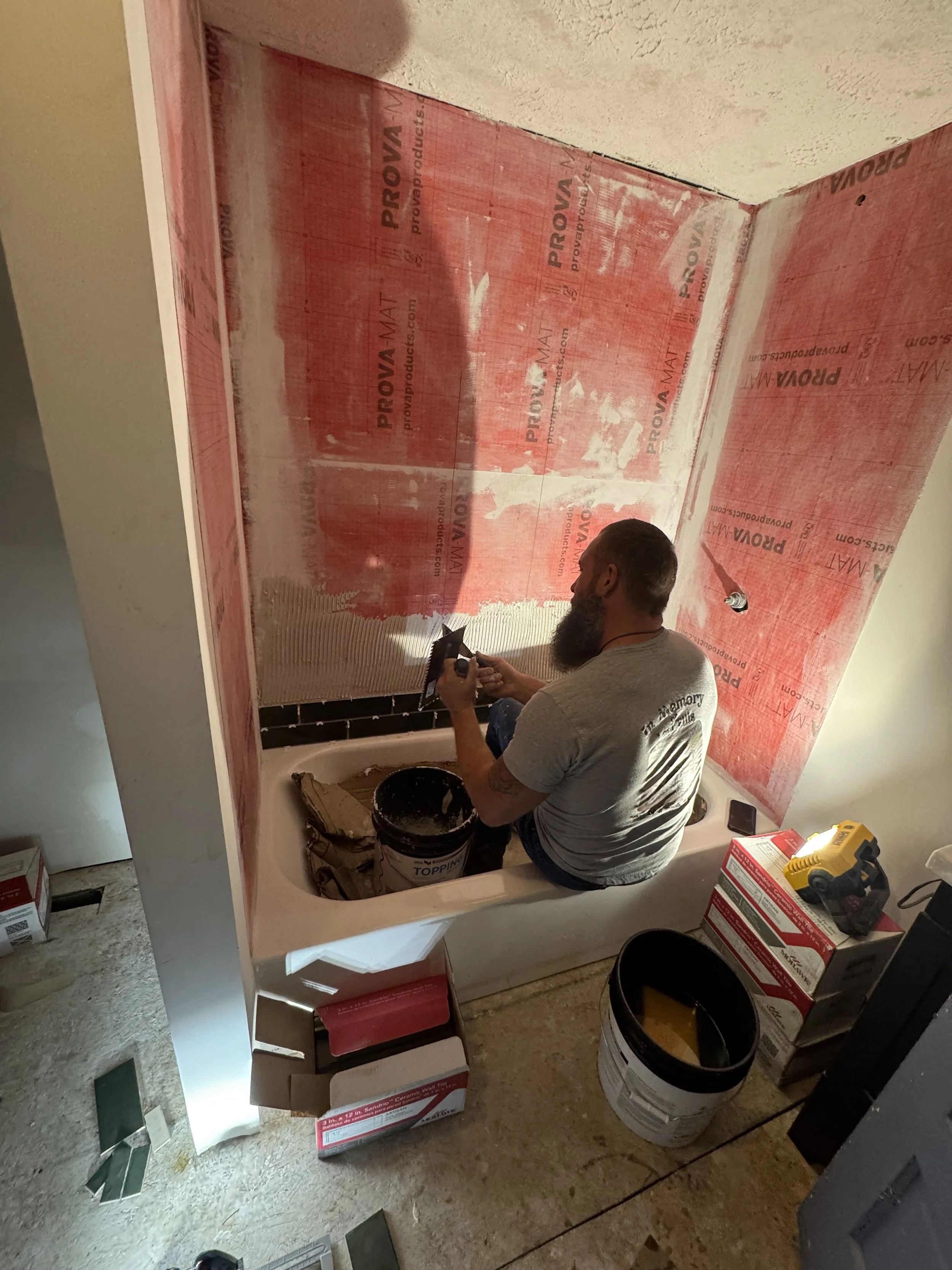 A man with a beard is kneeling inside a bathtub and using a power stapler. The bathtub is surrounded by boxes of construction materials, buckets, and tools. The wall above the bathtub is partially covered with red waterproofing material labeled 'PROV