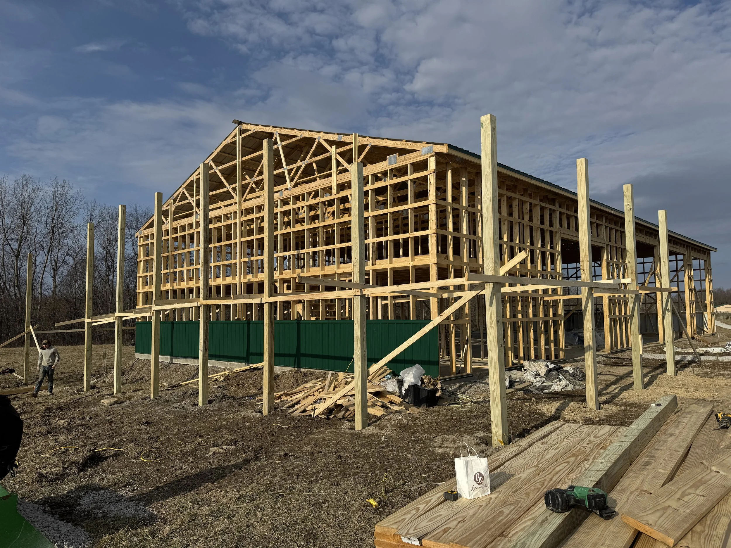 A house under construction with wooden framing, a sloped roof, and a worker walking on the site. Construction tools and materials are visible on the ground.