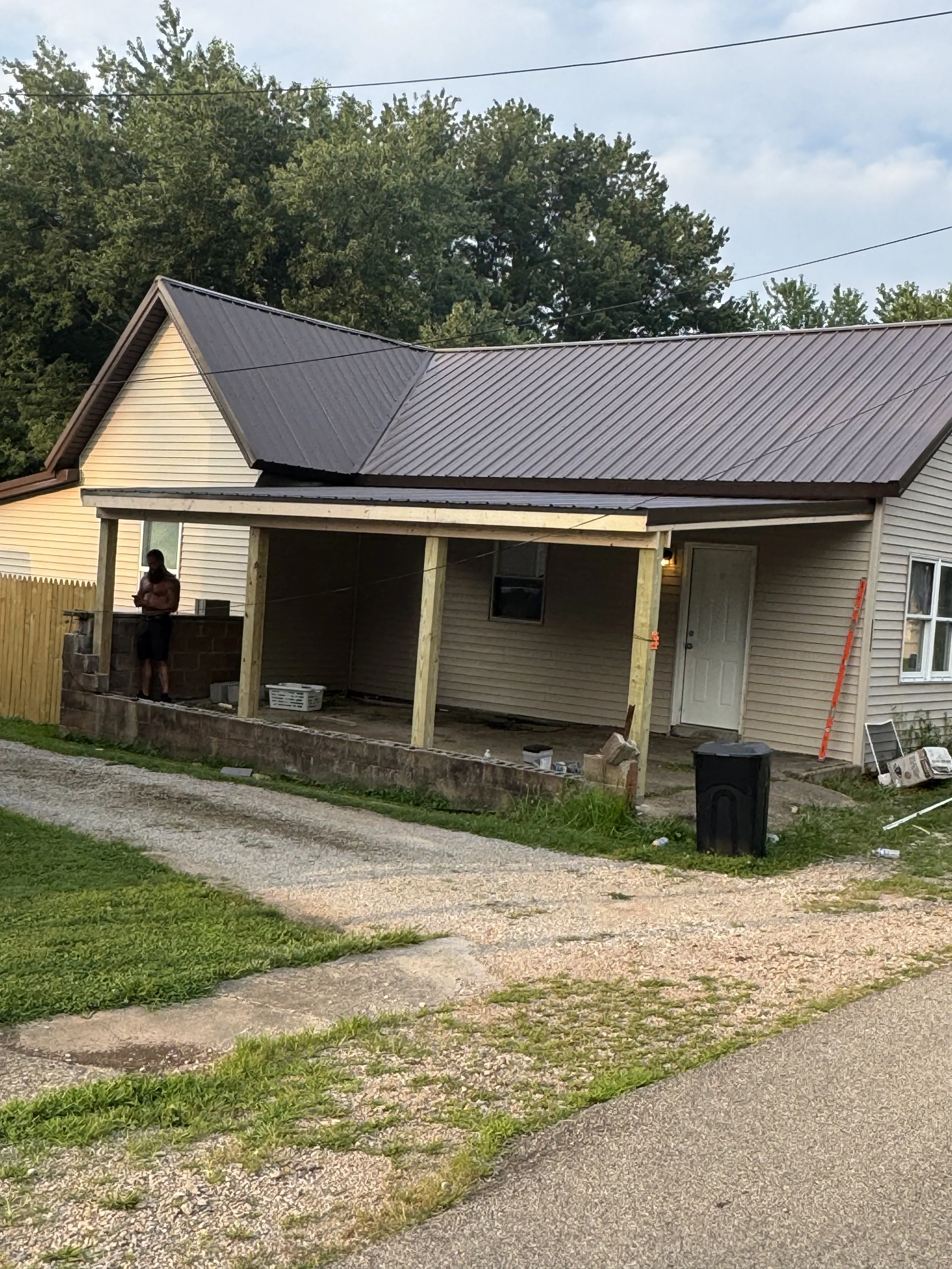 A house with a metal roof under construction, with a person standing on the porch, surrounded by green grass and trees in the background, under a partly cloudy sky.