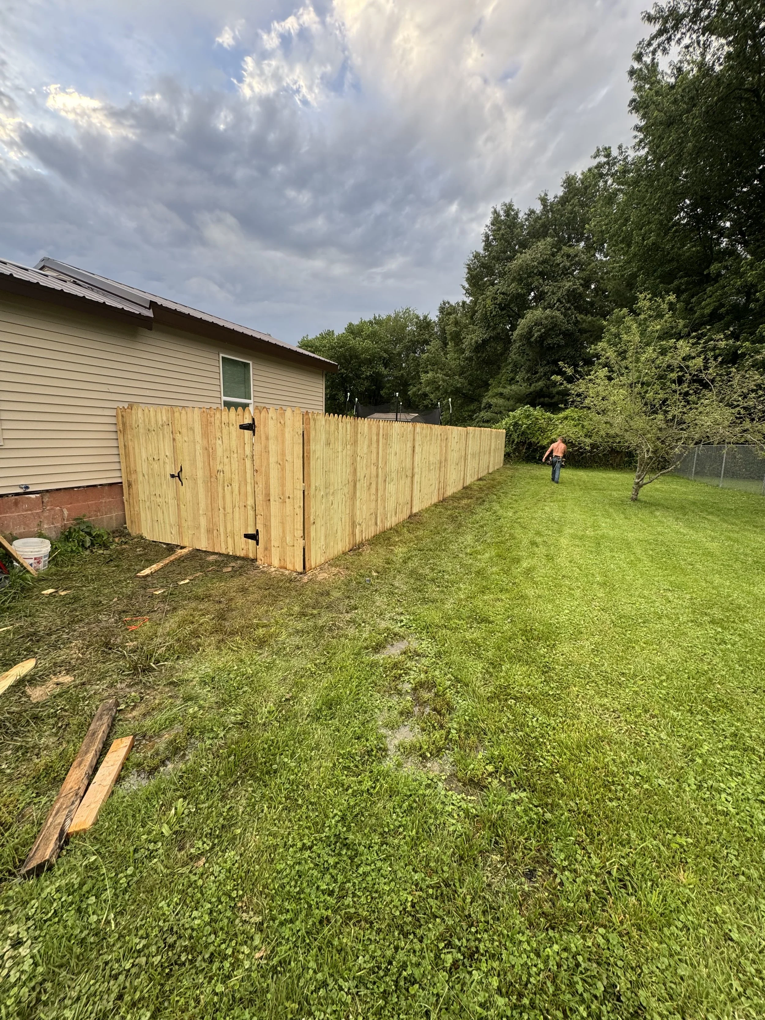 Residential backyard with newly built wooden fence, a house with beige siding, a tree, and a person in a pink shirt walking near the tree. Cloudy sky overhead.