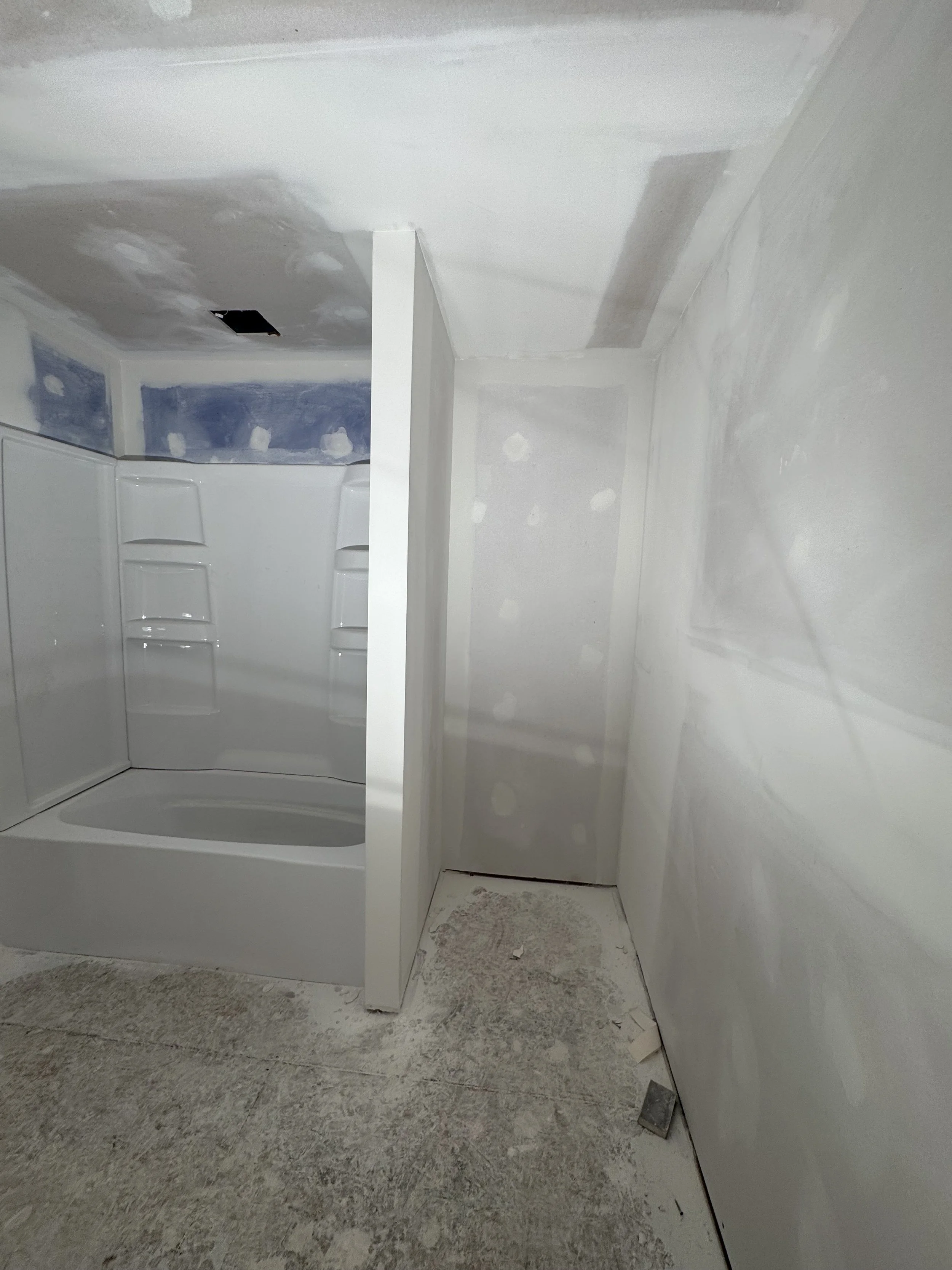 A bathroom under renovation with white walls, a corner shower stall, and overall unfinished space with visible dust and debris on the floor.