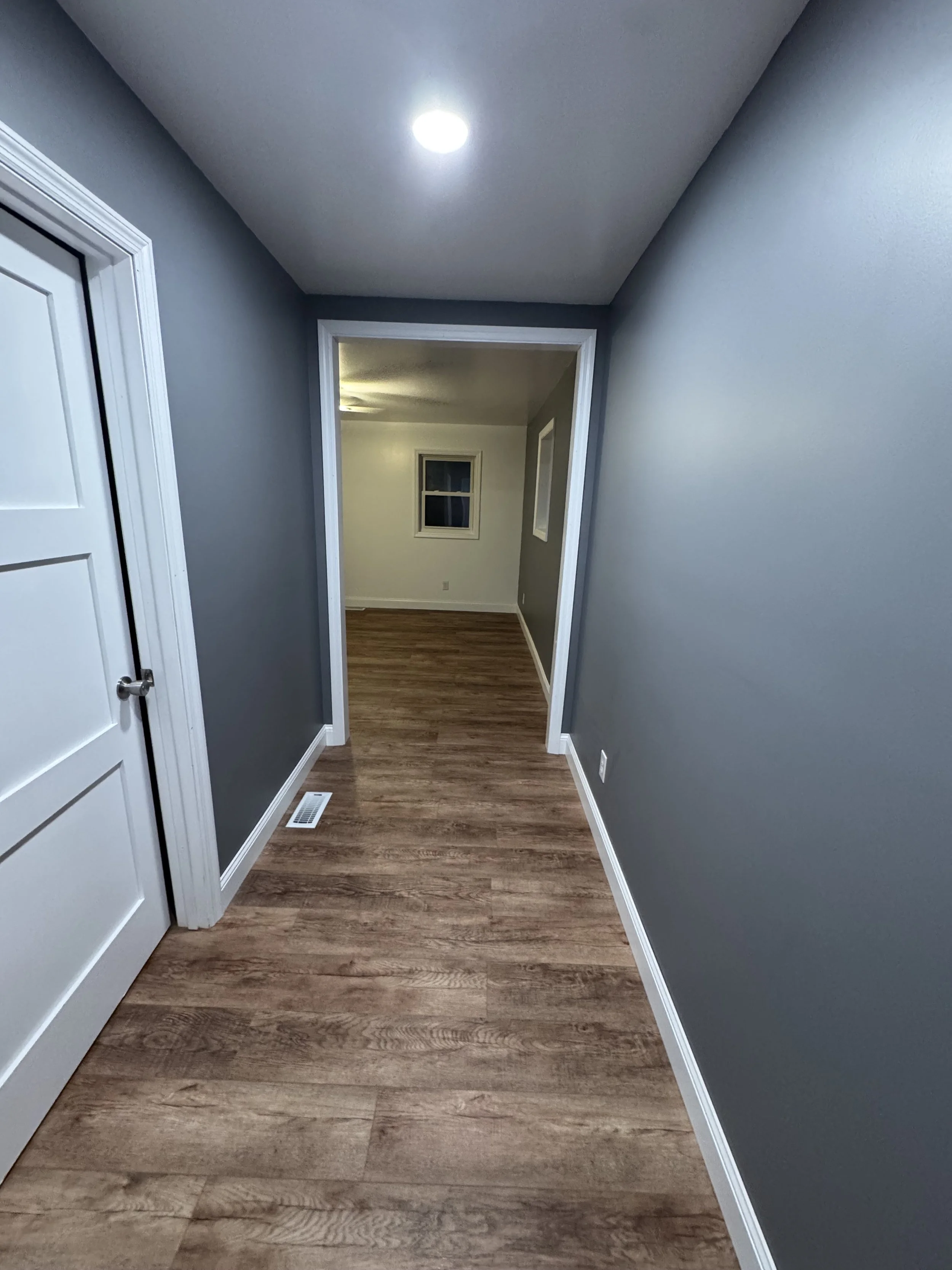 A hallway with gray walls and wood-patterned flooring, leading to a small room with a window and a ceiling light.