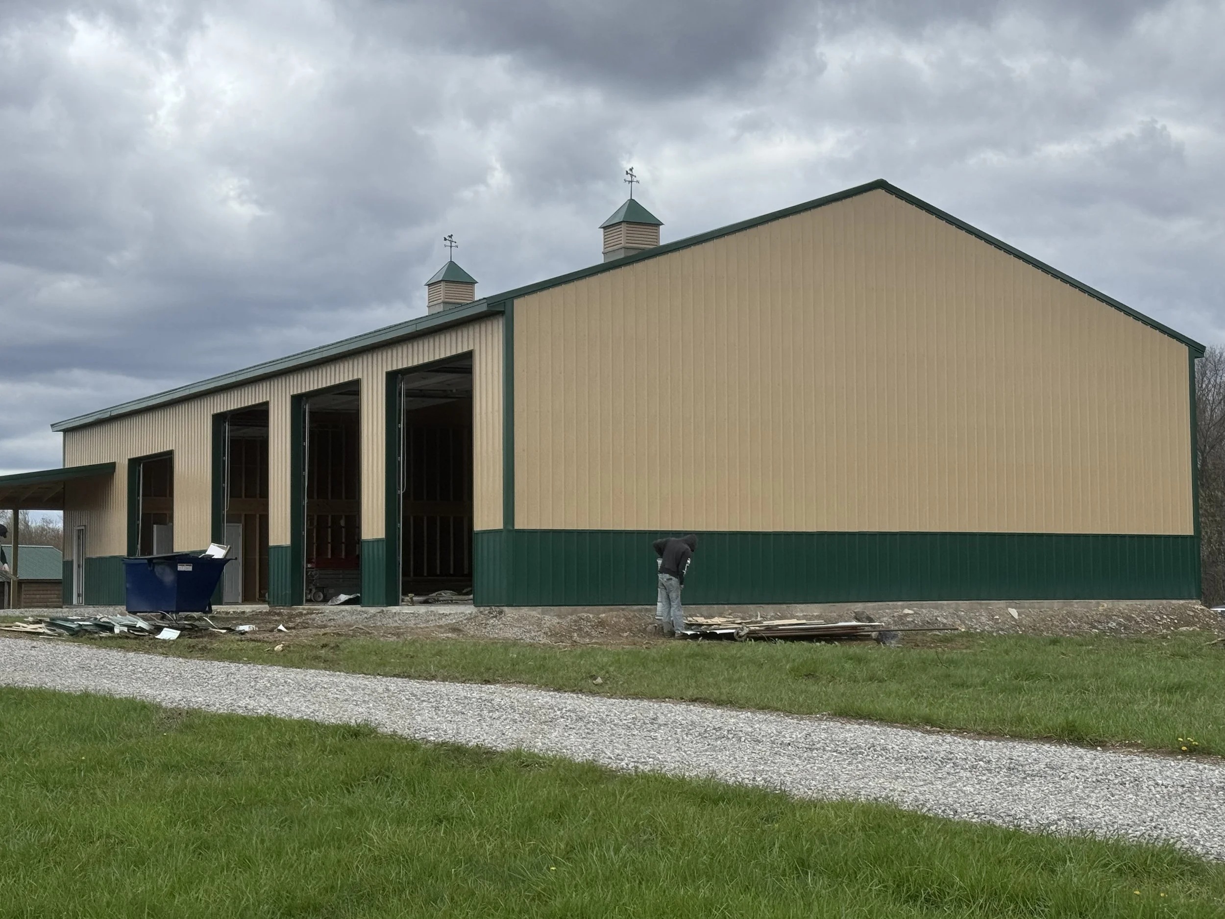 A building under construction with beige walls and green trim, featuring three small towers on the roof. The sky is cloudy. There is construction debris and a worker in the foreground.