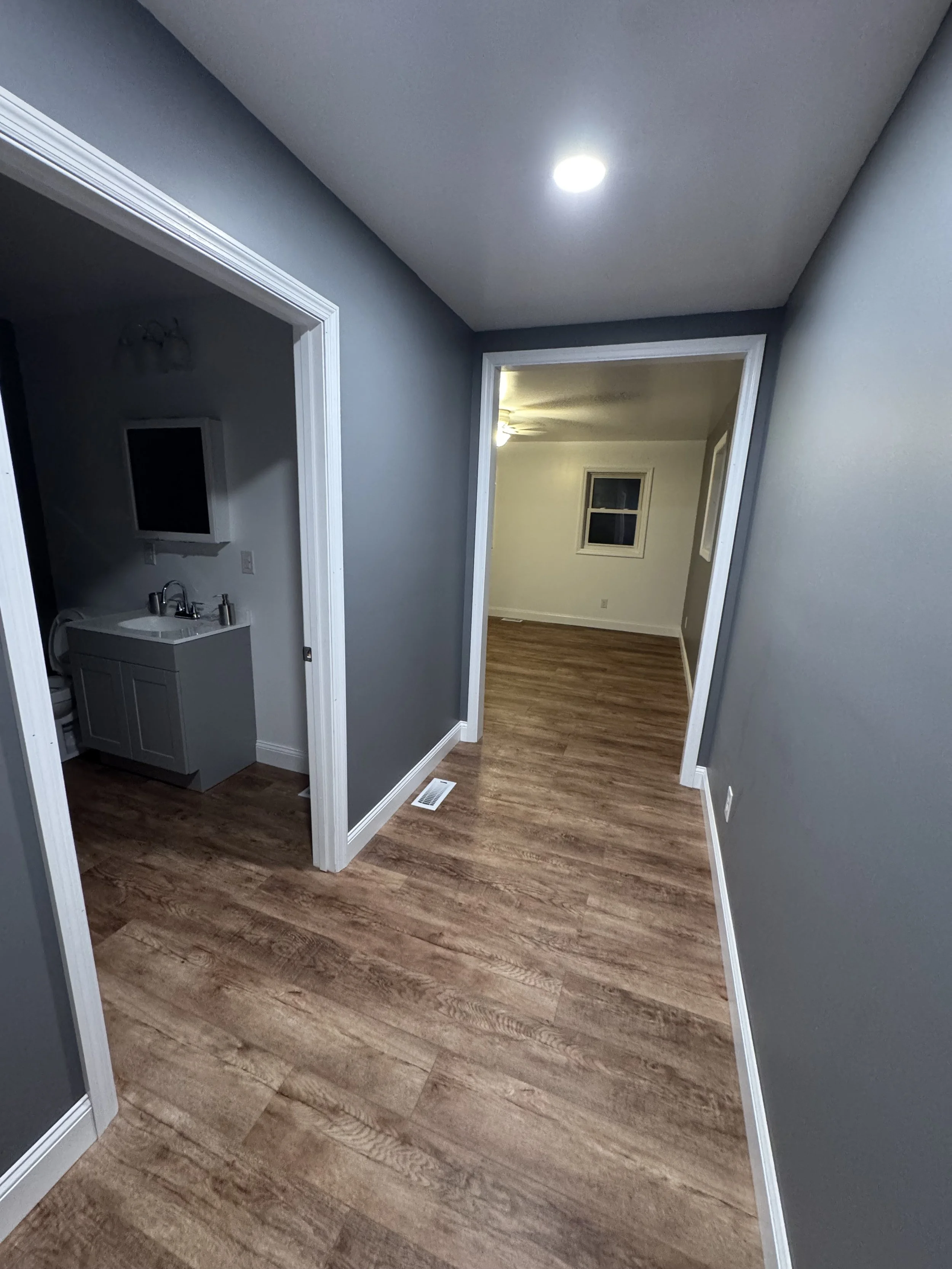 A hallway with hardwood floors, gray walls, white trim, and a ceiling light. An open doorway reveals a room with a ceiling fan and a window.