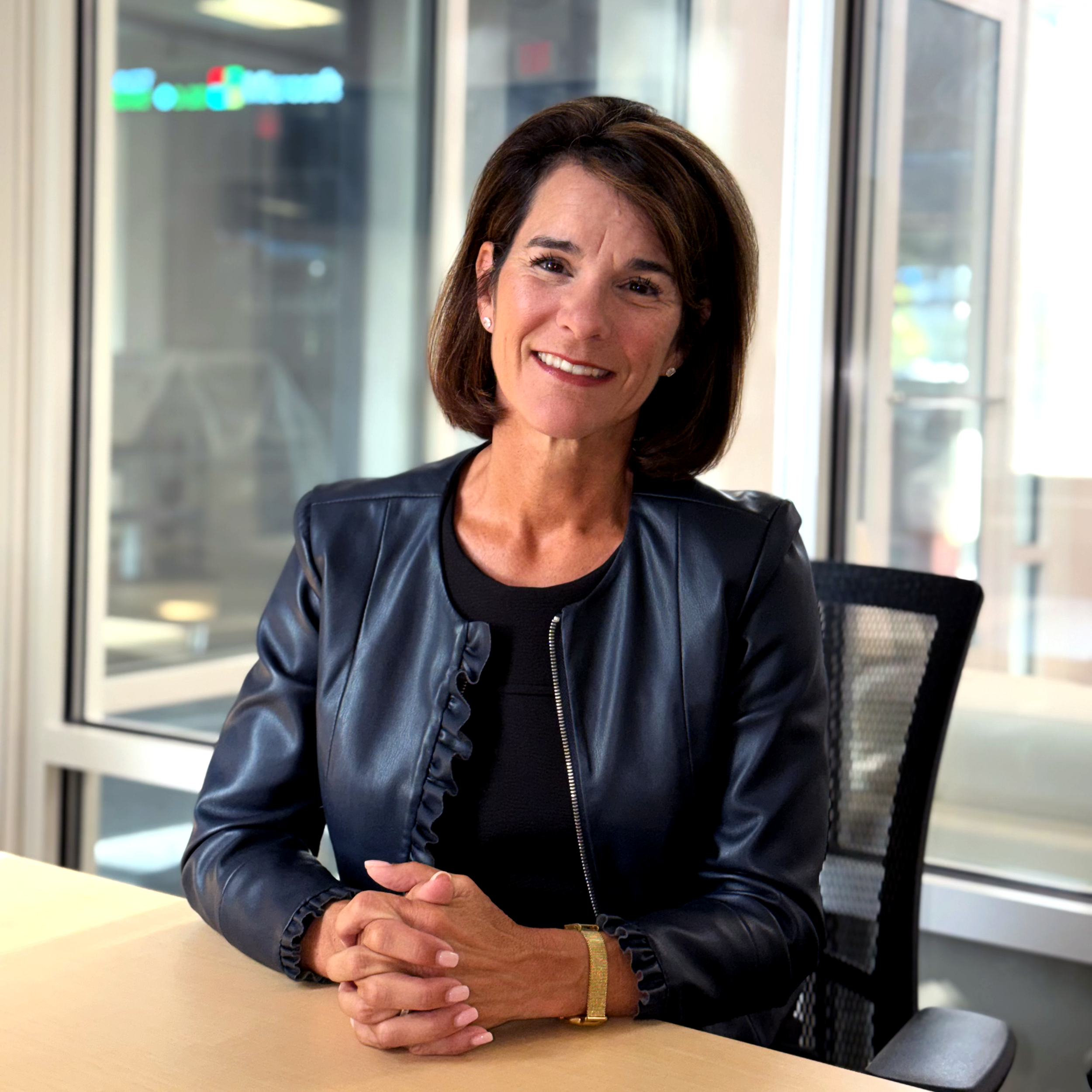 A woman with shoulder-length brown hair smiling in a professional setting, wearing a black leather jacket, sitting at a table with her hands clasped on top of it, with large windows and a modern office background. Dr. Jennifer Posa