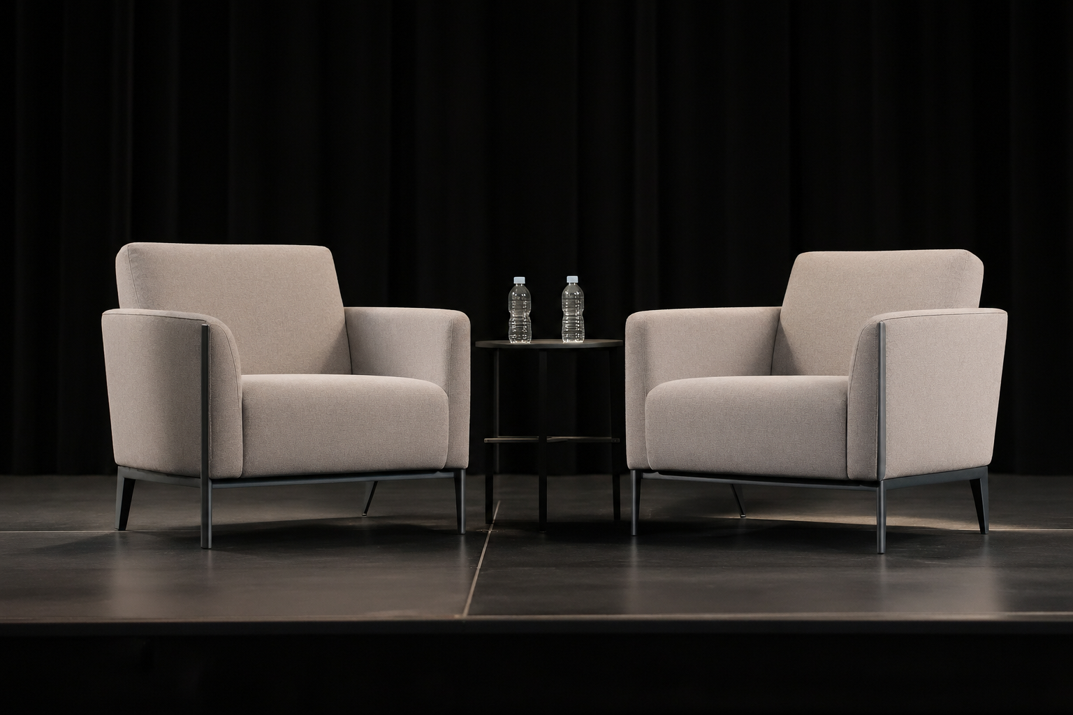 Two beige armchairs facing each other on a black stage with a small black table in between, holding two bottles of water, against a black curtain background.