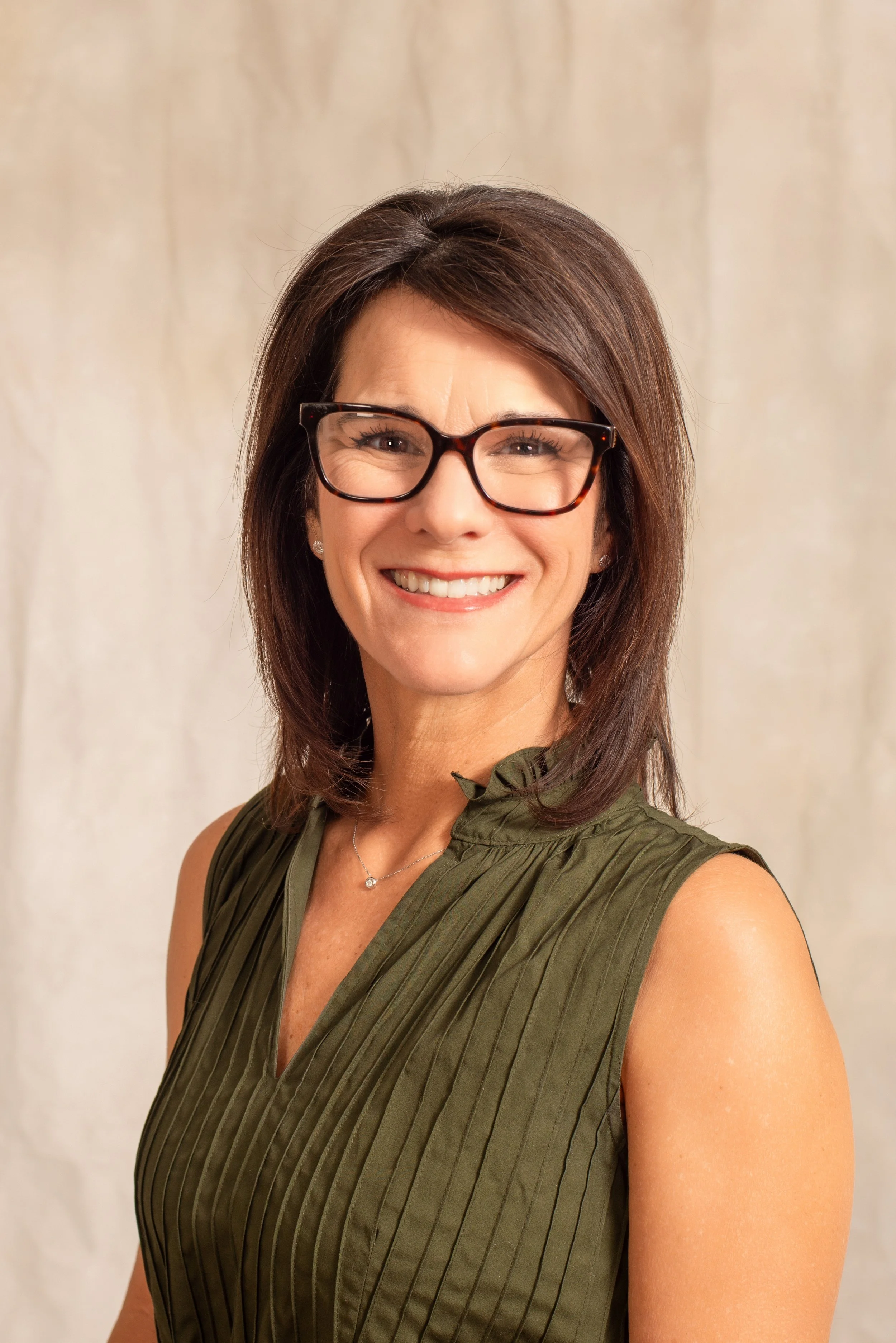 A smiling woman with brown hair, glasses, earrings, and a necklace, wearing an olive green sleeveless top with vertical pleats. Dr. Jennifer Posa
