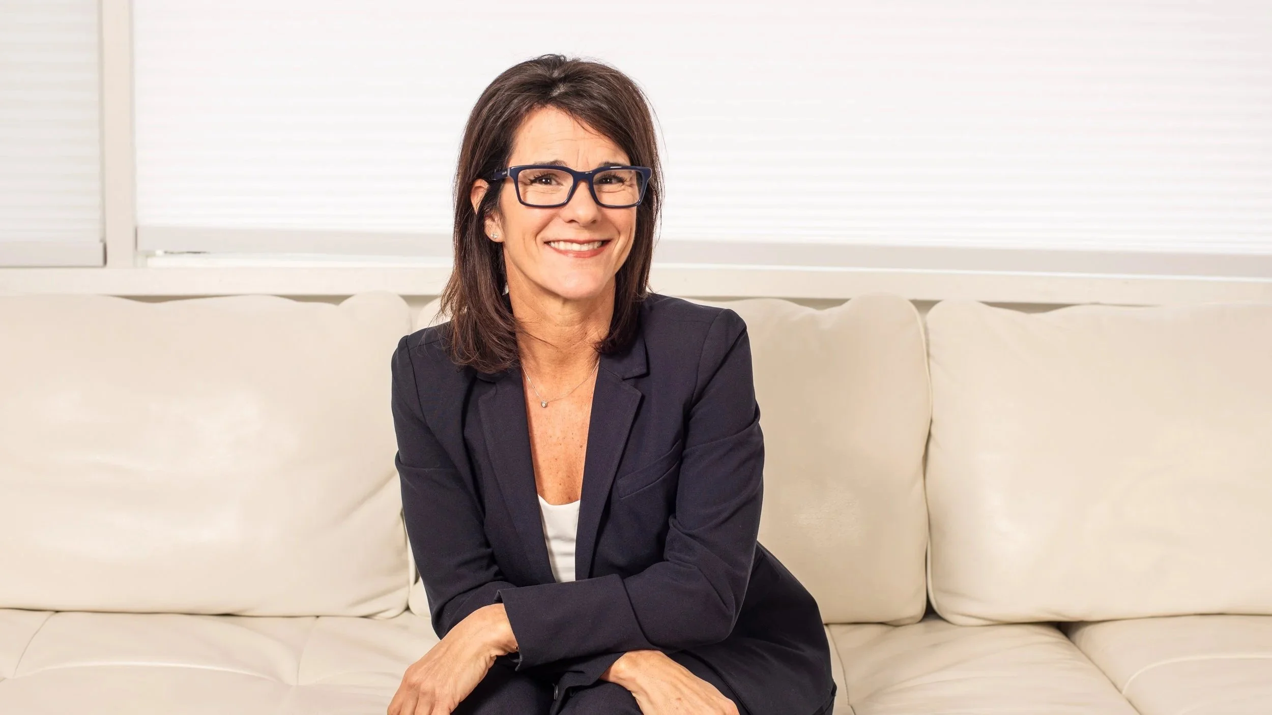 A middle-aged woman with shoulder-length brown hair, wearing glasses, a navy blazer, and a white shirt, sitting on a cream-colored sofa in front of a window with closed blinds, smiling at the camera.