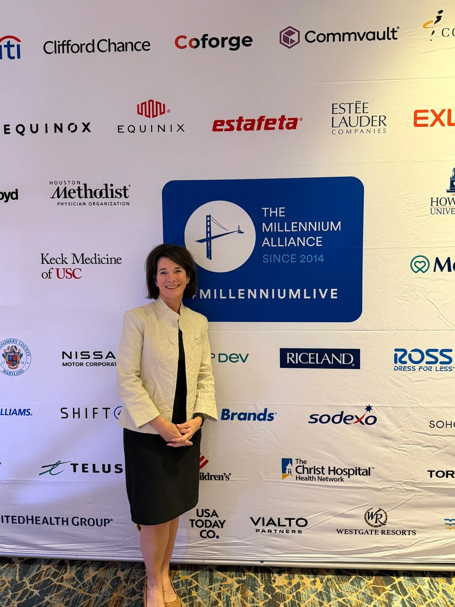 A woman standing in front of a backdrop with various logos of companies and organizations at an event, smiling and wearing a light-colored blazer and a black skirt. Dr. Jennifer Posa Keynote Event