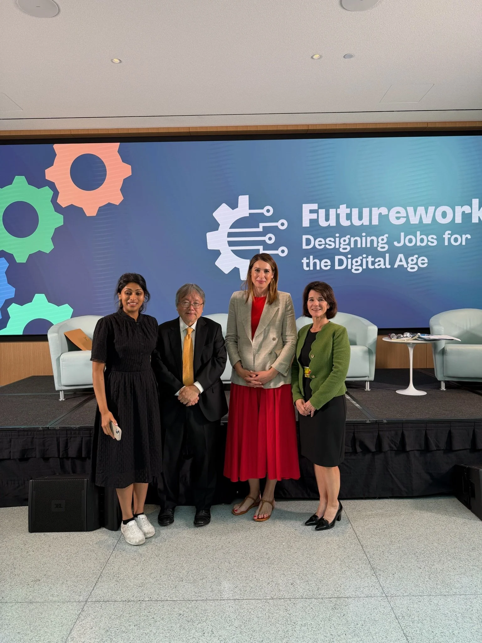 Four individuals standing together on a stage during a conference or event, with a large screen behind them displaying the title "Futurework: Designing Jobs for the Digital Age" and colorful gear icons. The group includes two women in formal attire, a man in a suit, and a woman in a blazer and skirt.