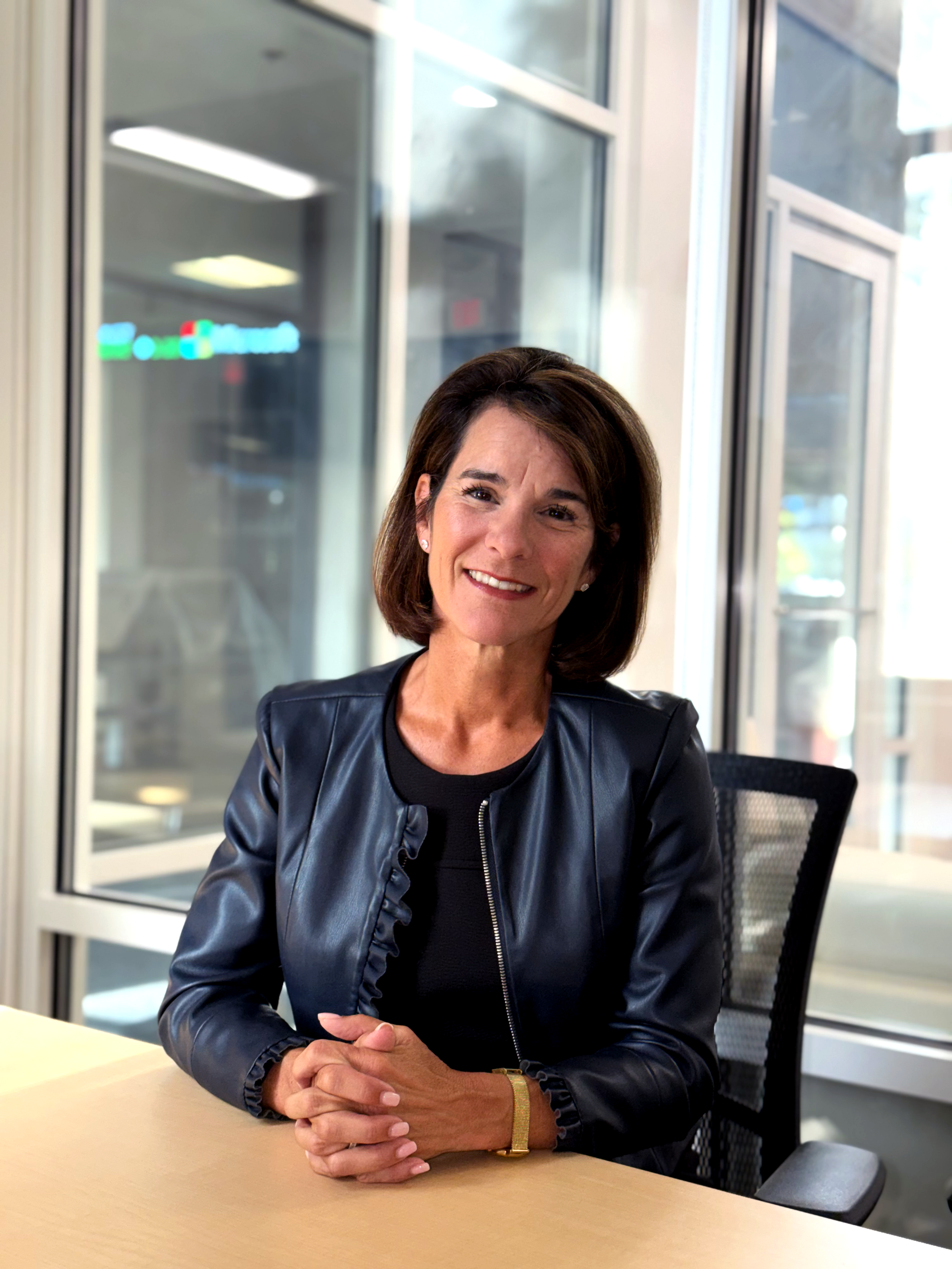A woman sitting at a conference table in an office setting, smiling, wearing a black leather jacket and a gold wristwatch, with large windows and a blurred company sign in the background. Dr. Jennifer Posa