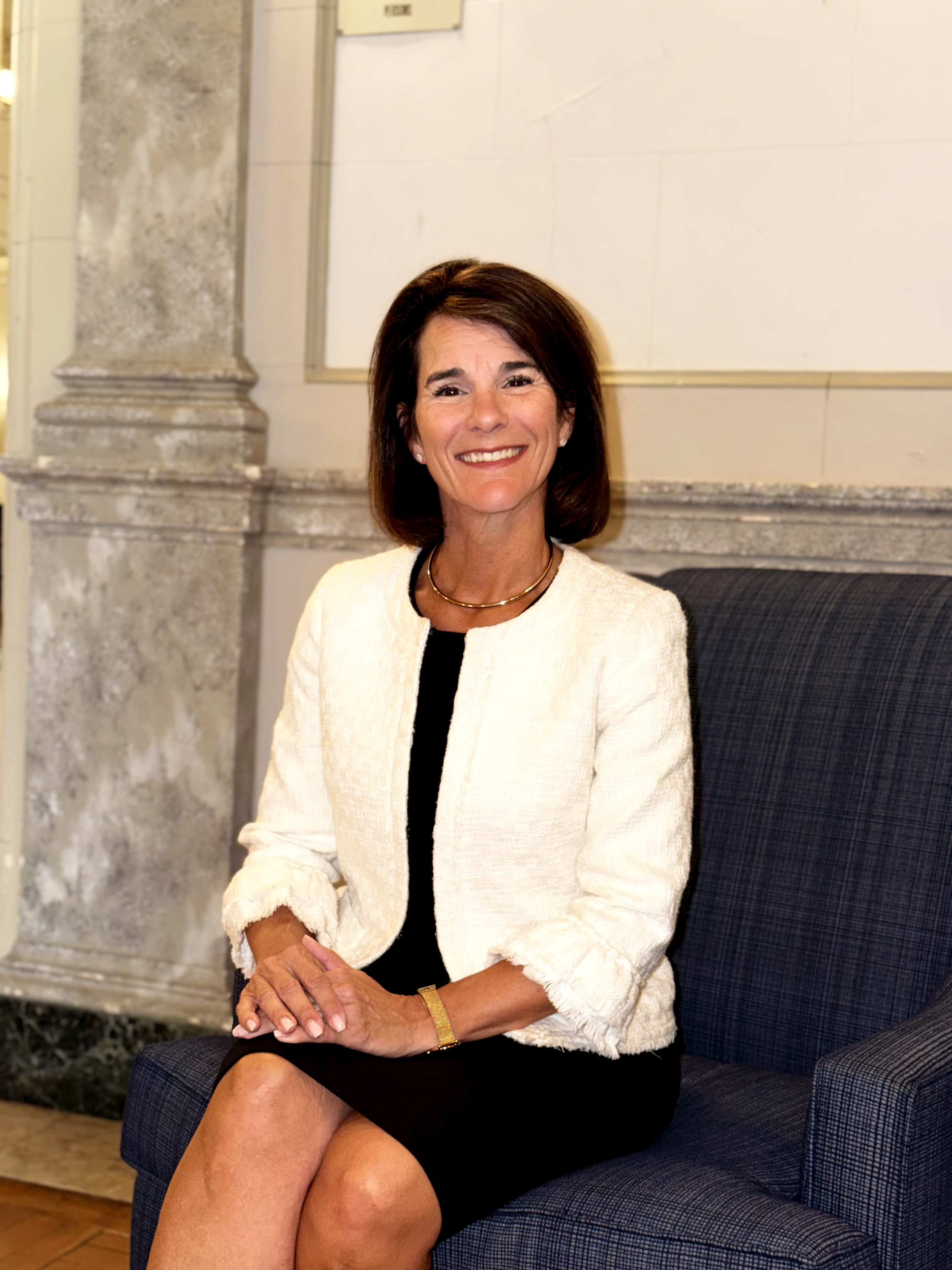 A woman with shoulder-length dark hair, wearing a white blazer with black details, a black dress, and gold jewelry, sitting on a blue armchair in a room with marble fireplace and beige walls. Dr. Jennifer Posa Photo