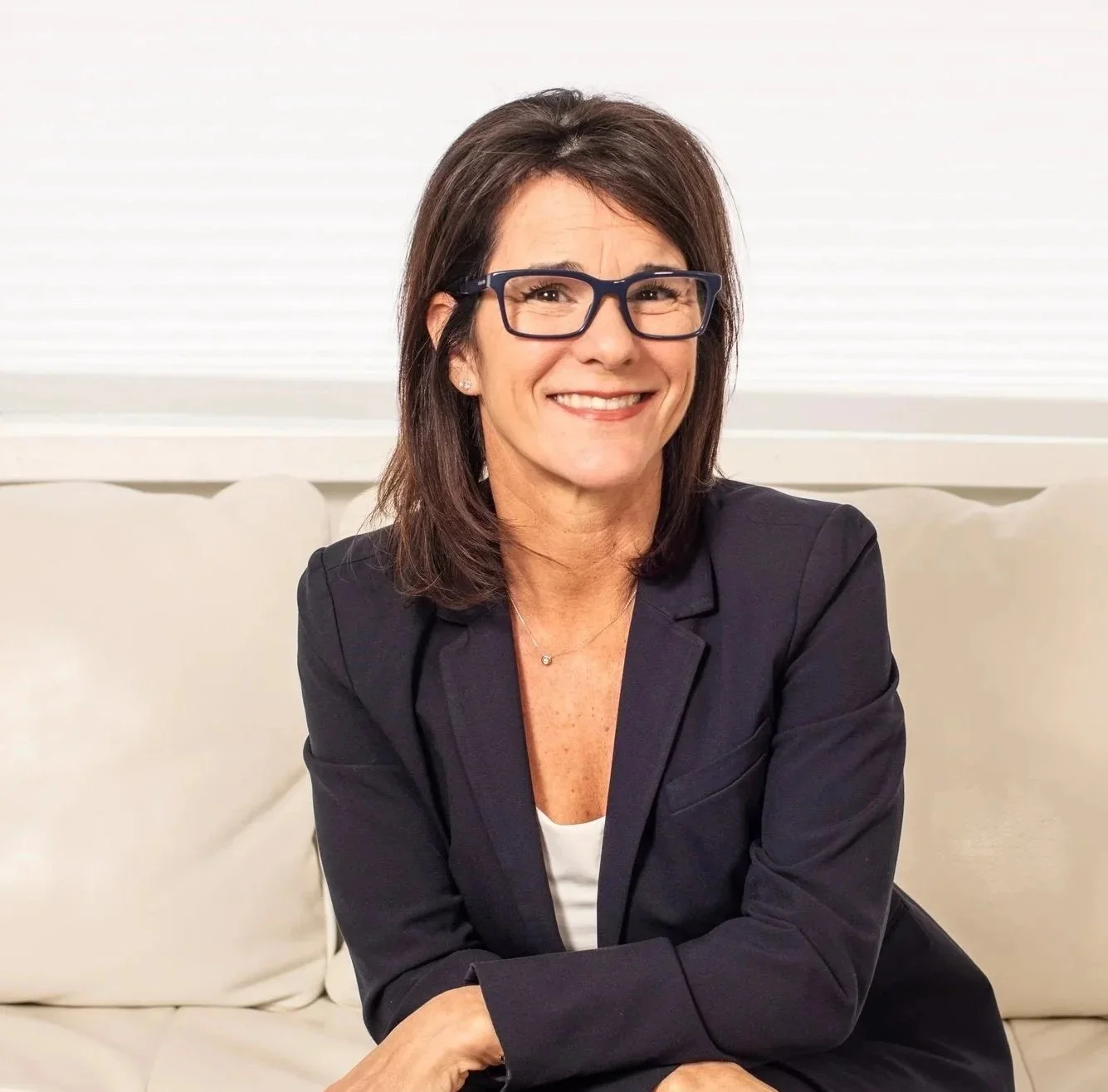 A woman with shoulder-length brown hair and glasses, wearing a black blazer and white top, seated on a cream sofa with a smile, in a well-lit room with white blinds. Dr. Jennifer Posa