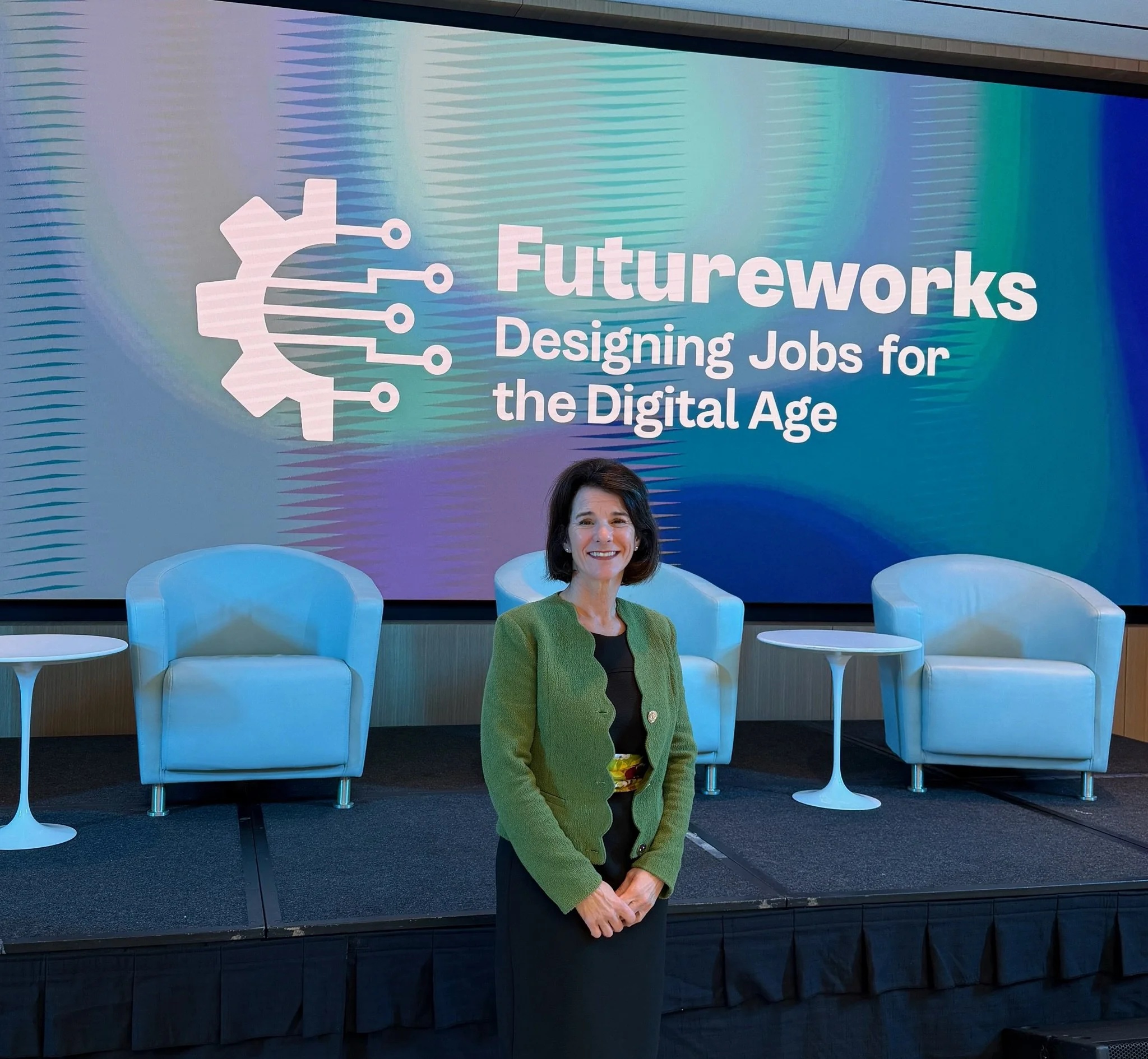 A woman with dark hair wearing a green blazer and black top, smiling, sitting on a white chair in front of a backdrop displaying the text 'Futureworks Designing Jobs for the Digital Age' and a graphic of a gear with circuit connections. Dr. Posa 