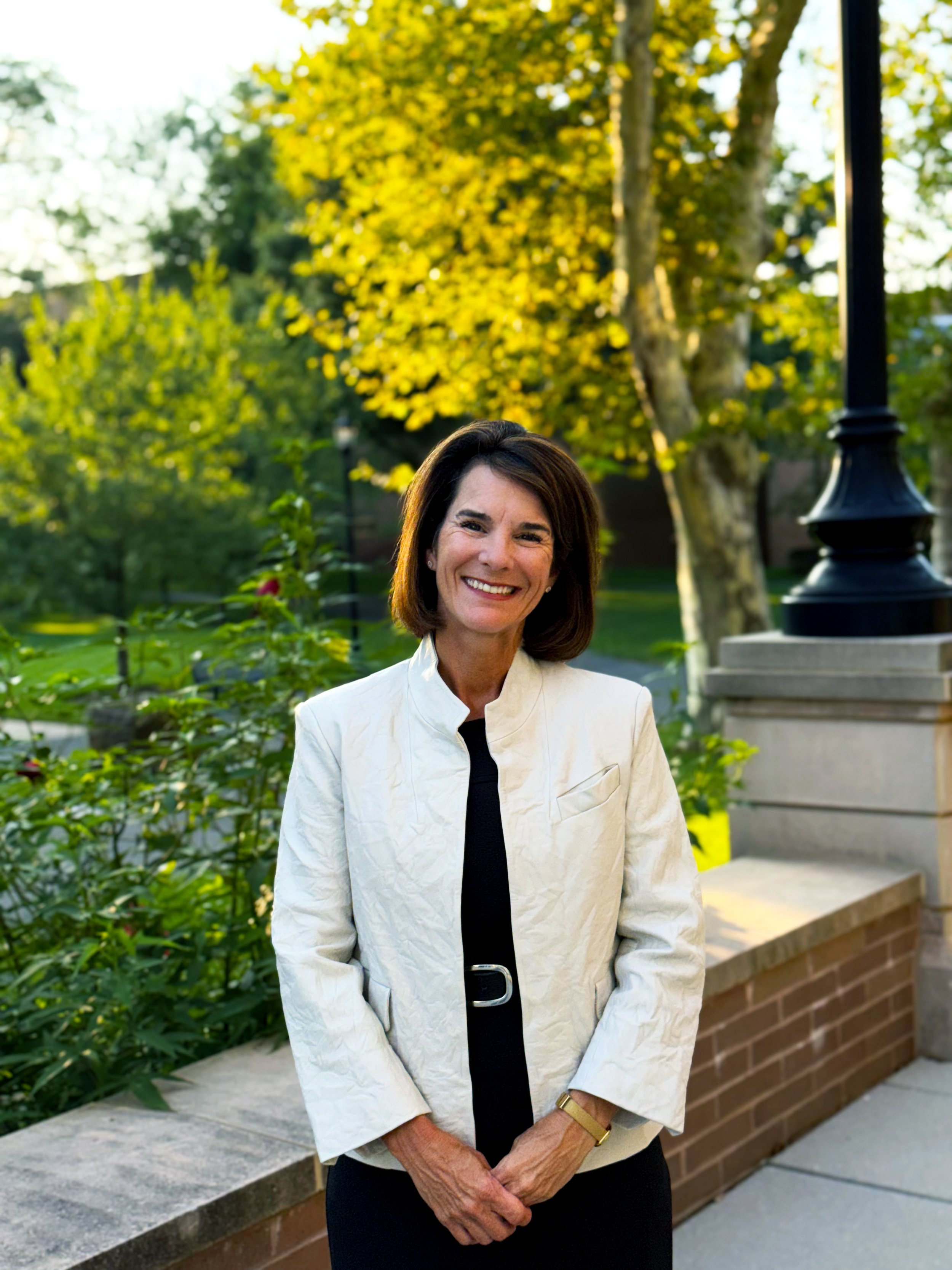 A smiling woman with shoulder-length dark hair, wearing a white jacket and black dress, standing outside near green trees and a brick wall during daytime. Dr. Jennifer Posa