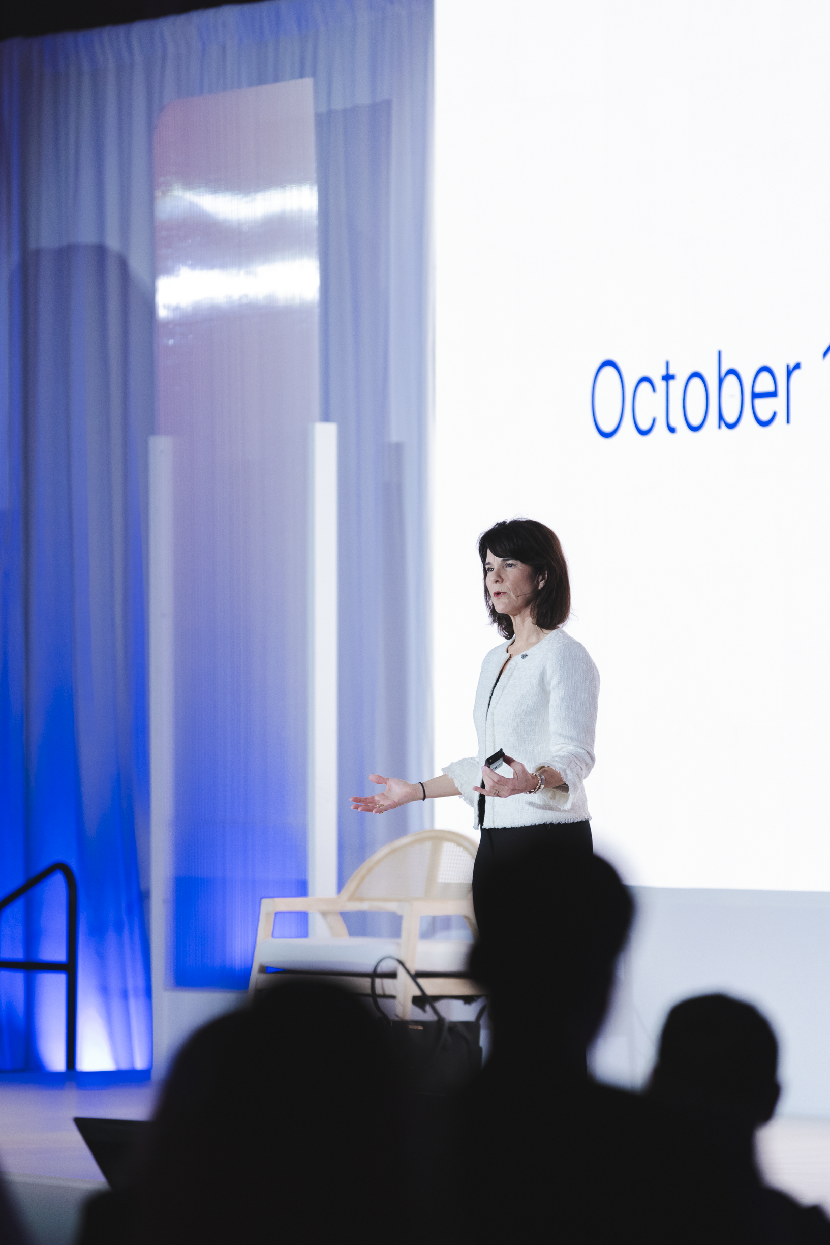 A woman giving a presentation on stage with a large screen behind her displaying the word 'October'. Dr. Jennifer Posa live keynote