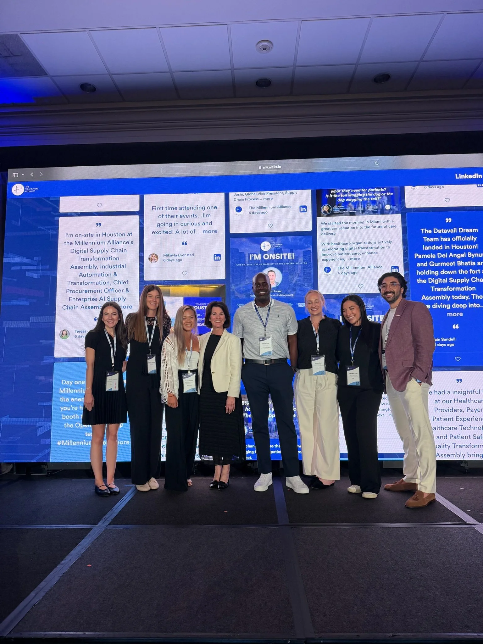 Group of seven people, four women and three men, standing together on a stage in front of a large digital display, smiling for the photo at a conference or event. Dr. Jennifer Posa Keynote event photo