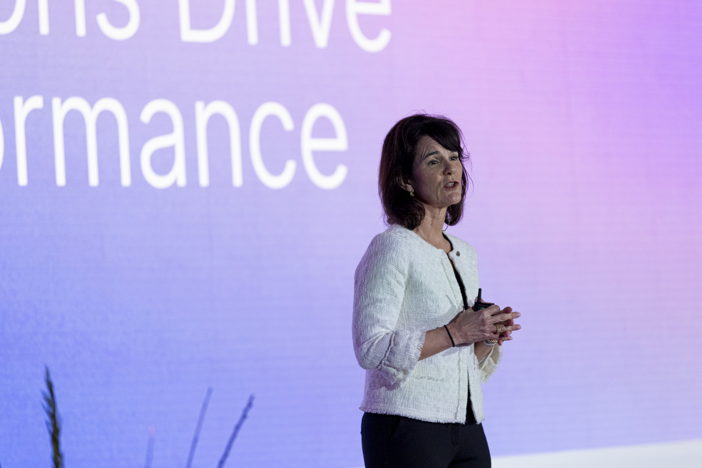 A woman is giving a presentation on a stage with a purple background. She is holding a microphone and appears to be speaking. The background has large, partially visible text that reads 'DRIVE' and 'rmance'. Dr. Jennifer Posa live keynote