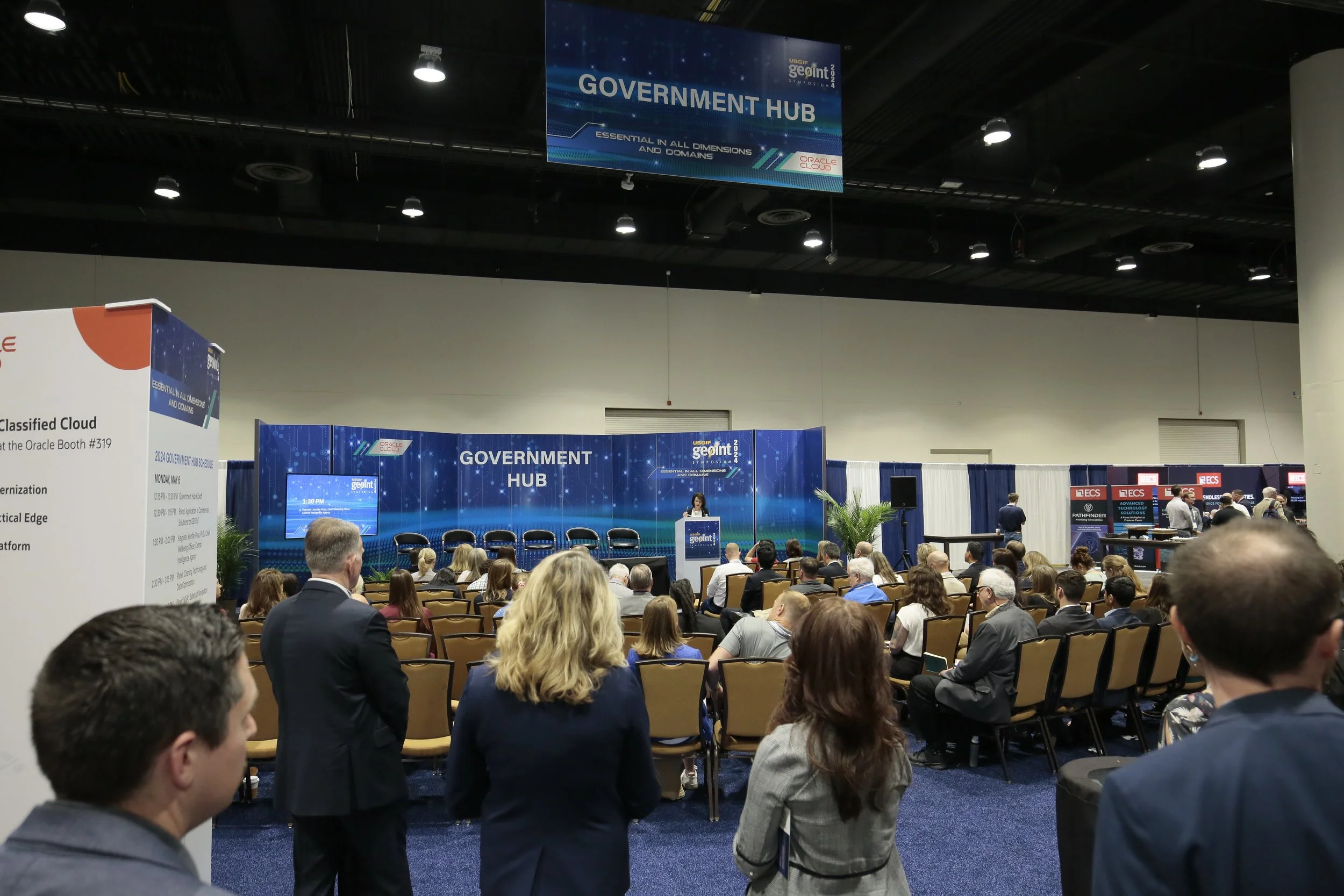A conference setting at a technology event with attendees seated facing a stage. The stage features a large blue backdrop with the words 'GOVERNMENT HUB' and a speaker presenting at a podium. Overhead digital screens display event branding and sponsor information. The room is filled with people, some standing and some sitting, with a mix of business attire. There are exhibition booths along the side with company logos.