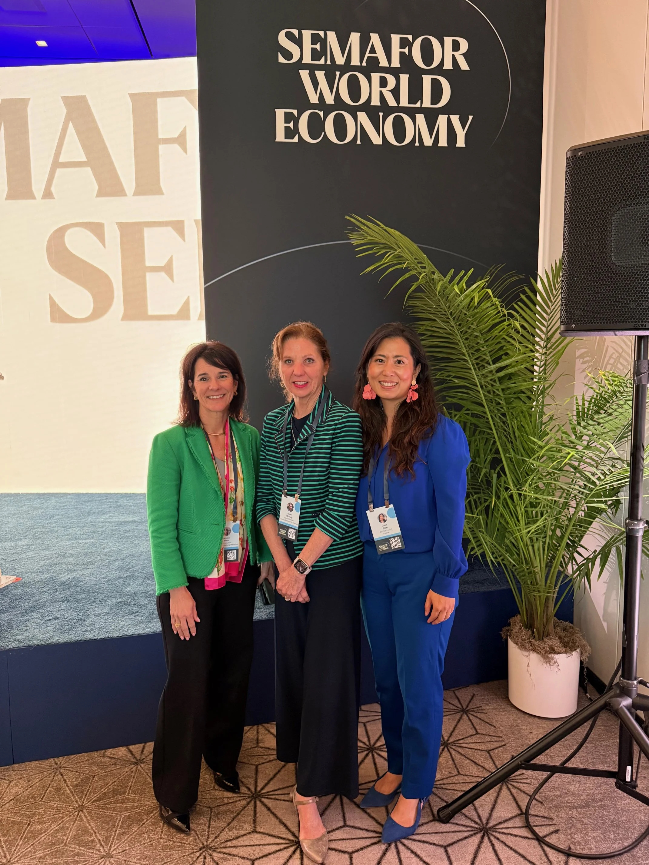 Three women standing together at a conference or event, smiling, with a backdrop that says "SEMAFOR WORLD ECONOMY." They are wearing conference badges, and there is a large potted plant next to them. Dr. Jennifer Posa with others at Event