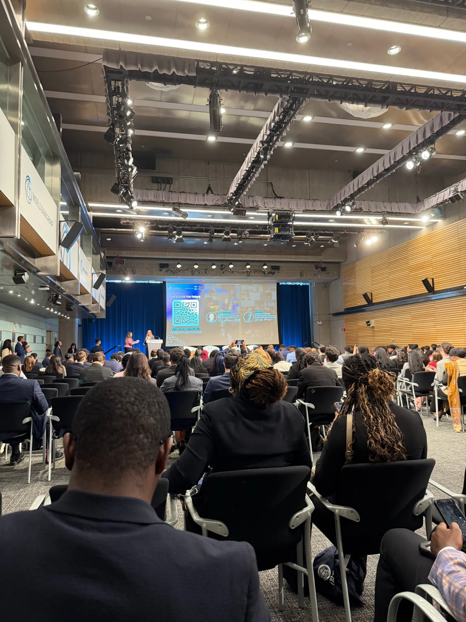 A large conference or event hall filled with people seated and listening to a presentation. Two speakers stand at a podium on a stage with a large screen displaying a QR code and promotional content. The hall has a high ceiling with various lighting and sound equipment.