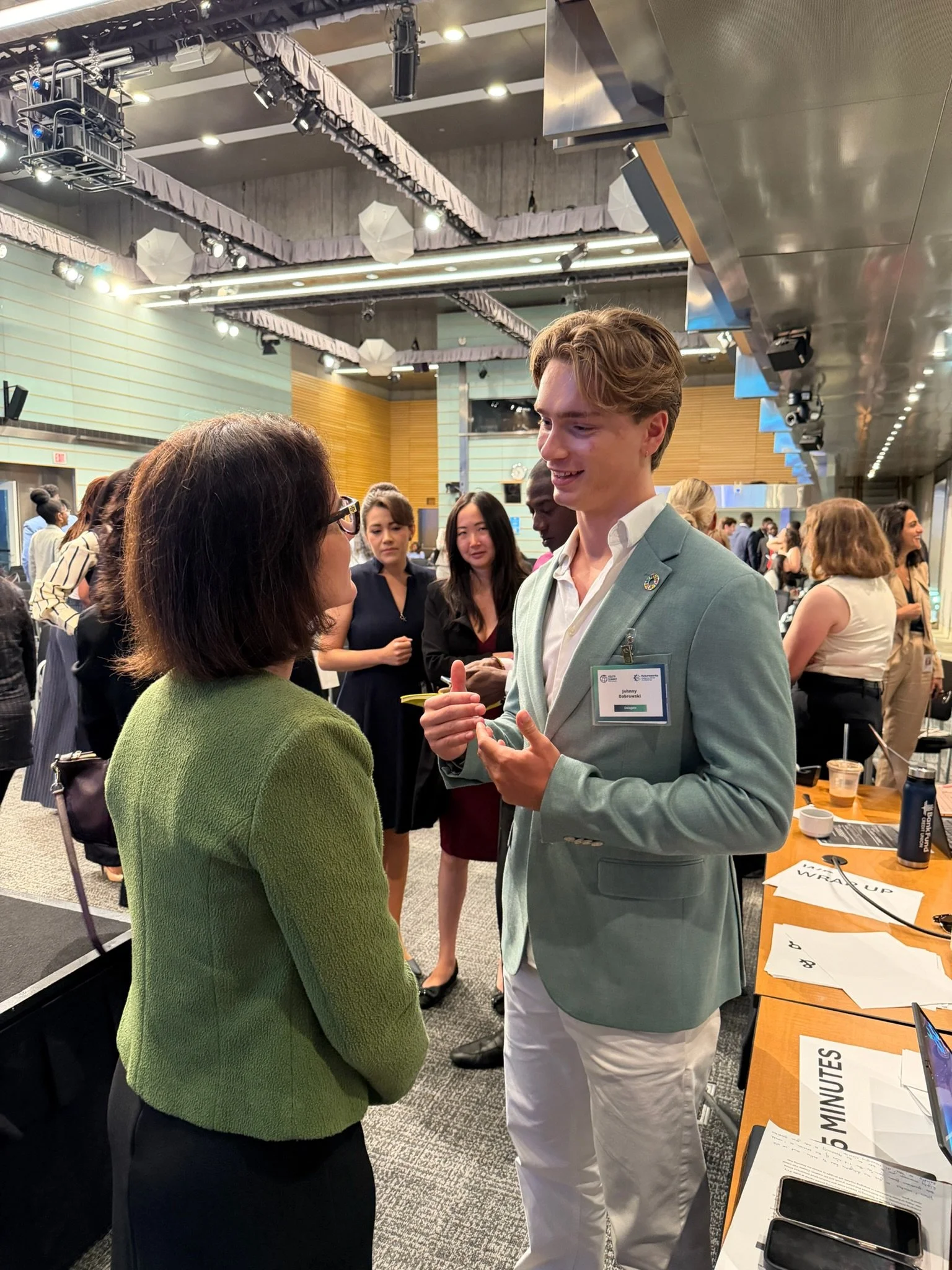A young man in a light blue blazer and white pants is talking to a woman with glasses and a green sweater; they are at a busy event or conference with other people in the background. Dr. Jennifer Posa post-keynote conversations