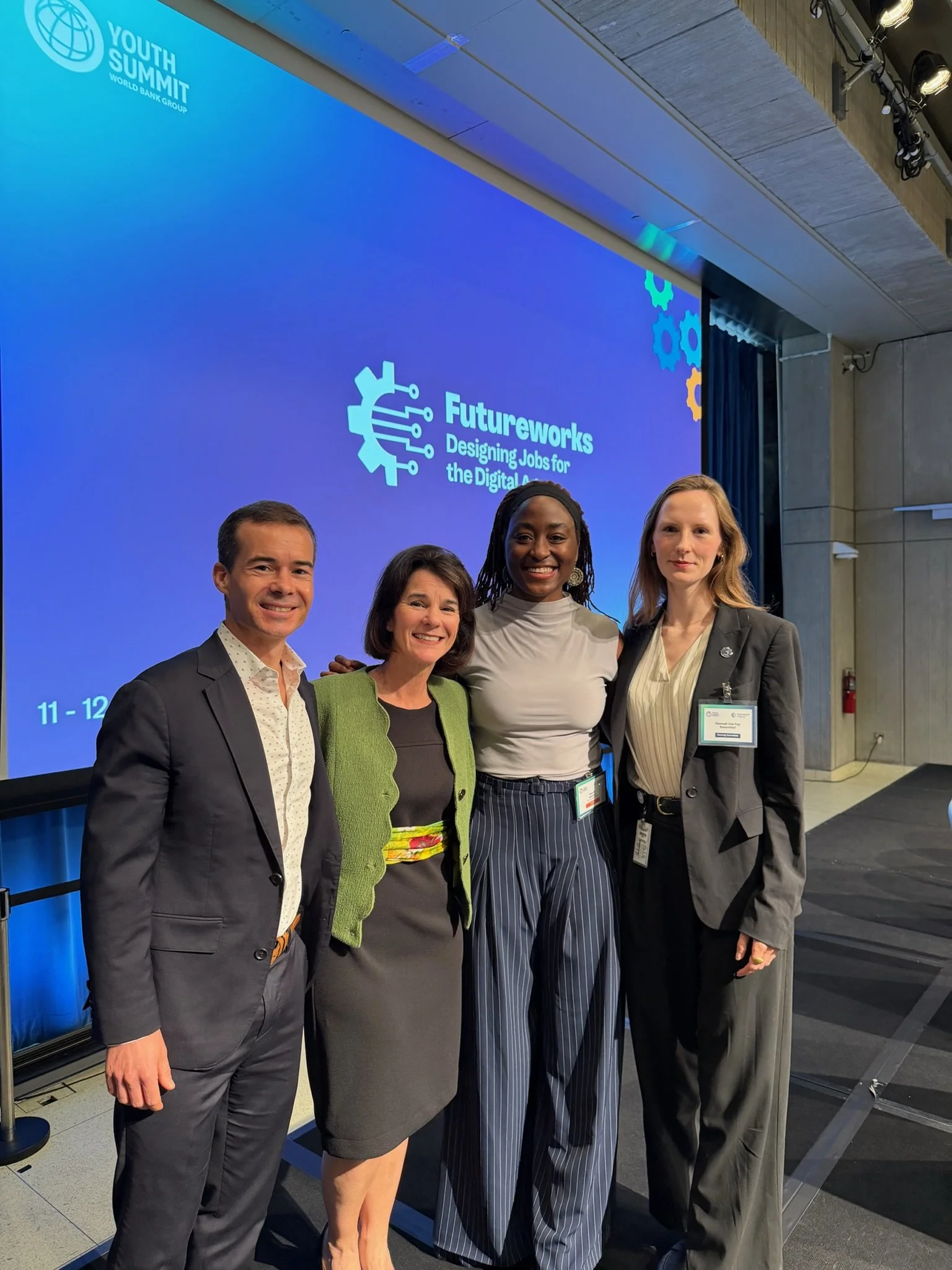 Group of four people standing together in front of a blue screen at the Youth Summit organized by World Bank Group. The screen displays the event logo and the theme 'Futureworks: Designing Jobs for the Digital Age.' The people are dressed in business attire, smiling, and appear to be at a professional conference.