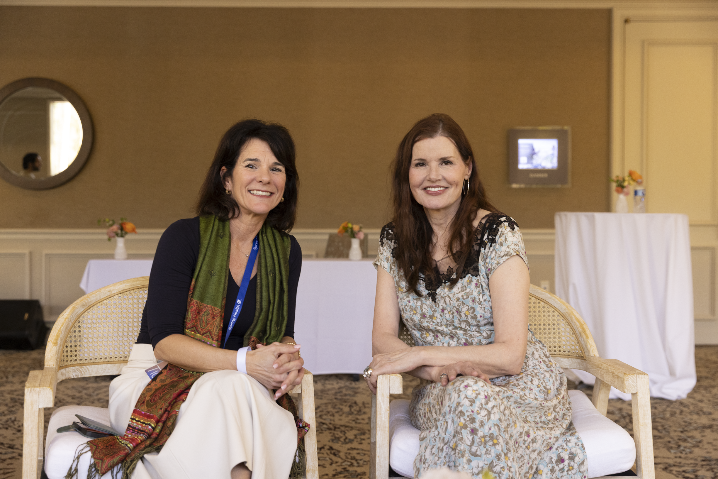 Two women sitting on wooden chairs at a table, smiling at the camera in a conference room decorated with small vases of flowers on tables and a wall with a mirror and a monitor. Dr. Jennifer Posa event photo