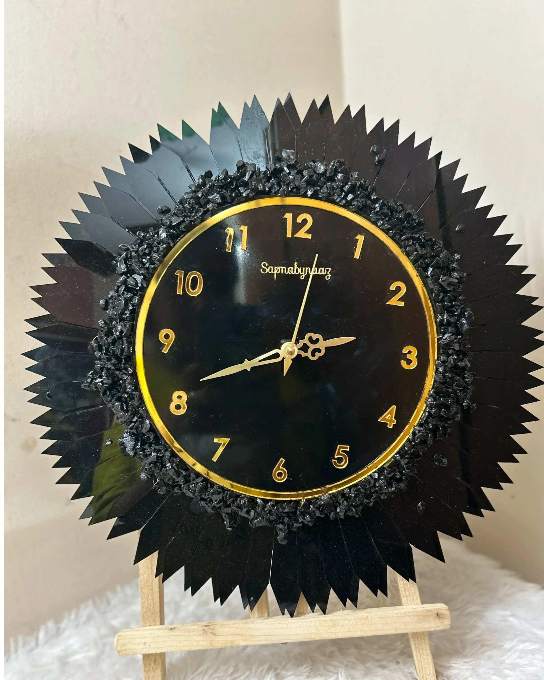 A decorative black and gold clock on a wooden easel, with a spiked outer rim and black crystals around the face, displaying the time as approximately 2:45.