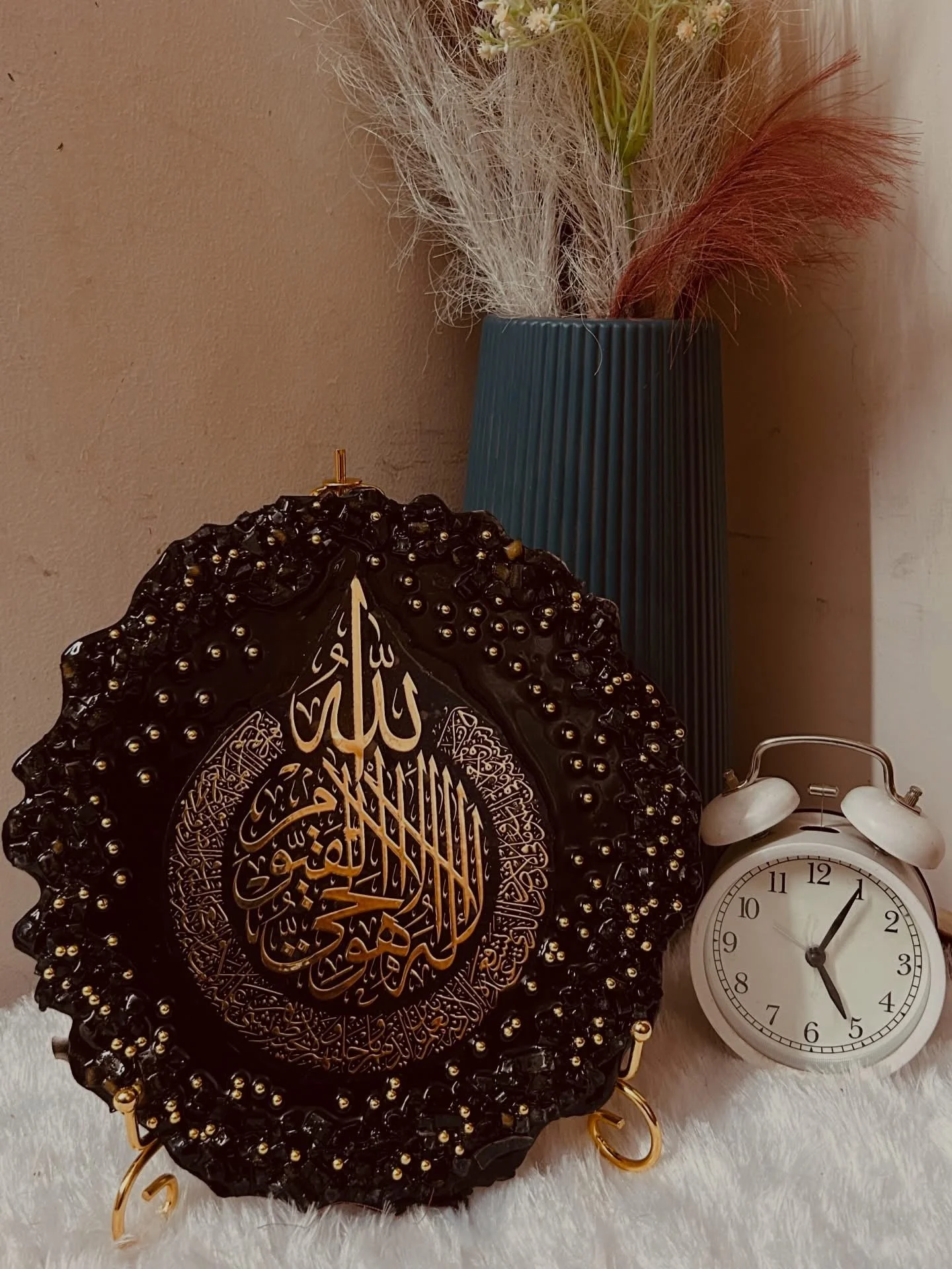 Decorative black and gold Islamic art piece with Arabic calligraphy, a white alarm clock showing 5:02, a tall teal vase with dried flowers, and a white wall clock, all arranged on a furry surface.