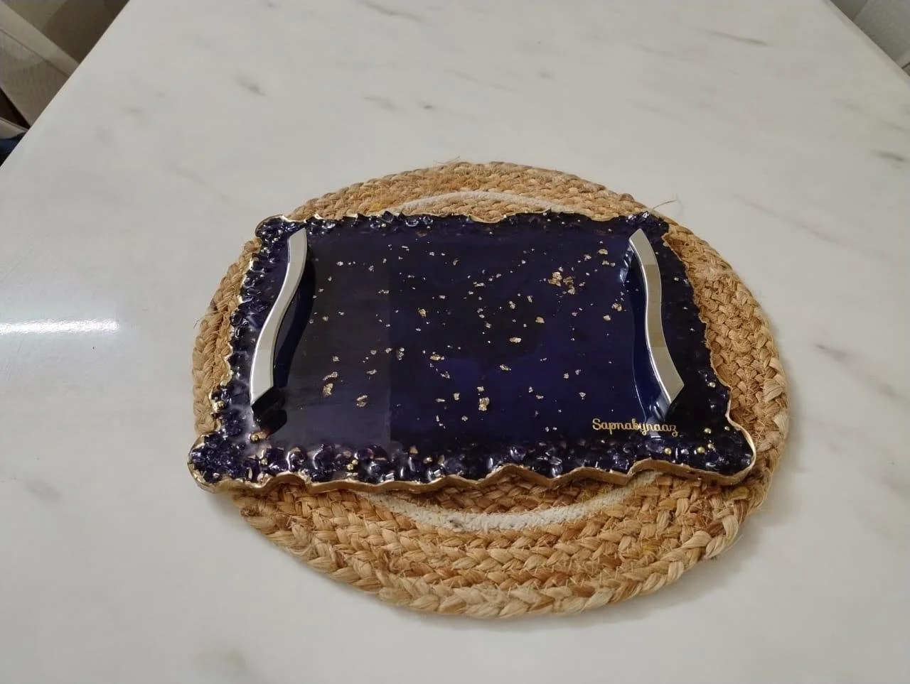 Decorative serving tray with a black glossy surface, gold speckles, and silver handles, placed on a round woven placemat on a white marble table.