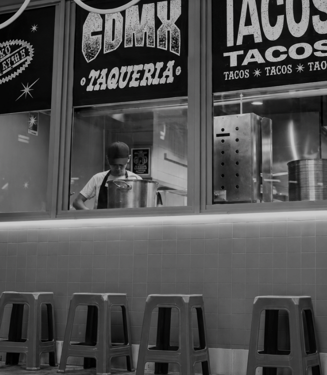 Inside a taco restaurant, a person in a baseball cap and apron is cooking behind a glass window with signs that read 'CDMX Taqueria' and 'Tacos.' In the foreground, four empty stools are aligned against a tiled wall.