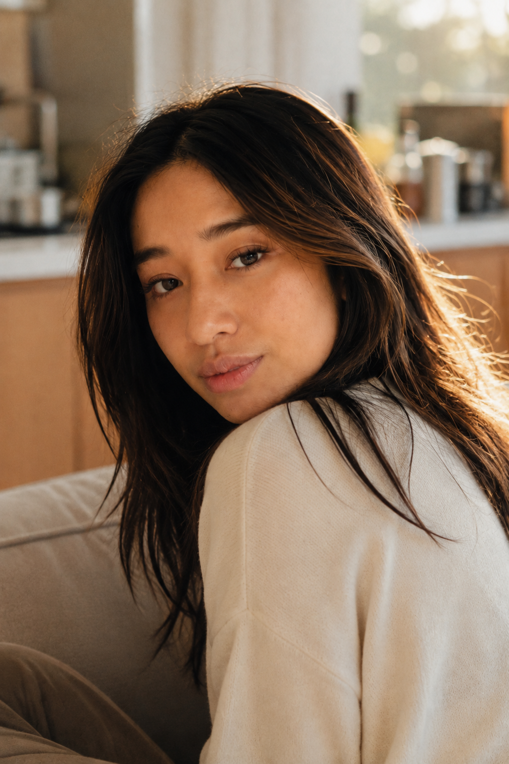 A young woman with long dark hair and fair skin looking at the camera with a relaxed expression, sitting on a couch in a warmly lit room.