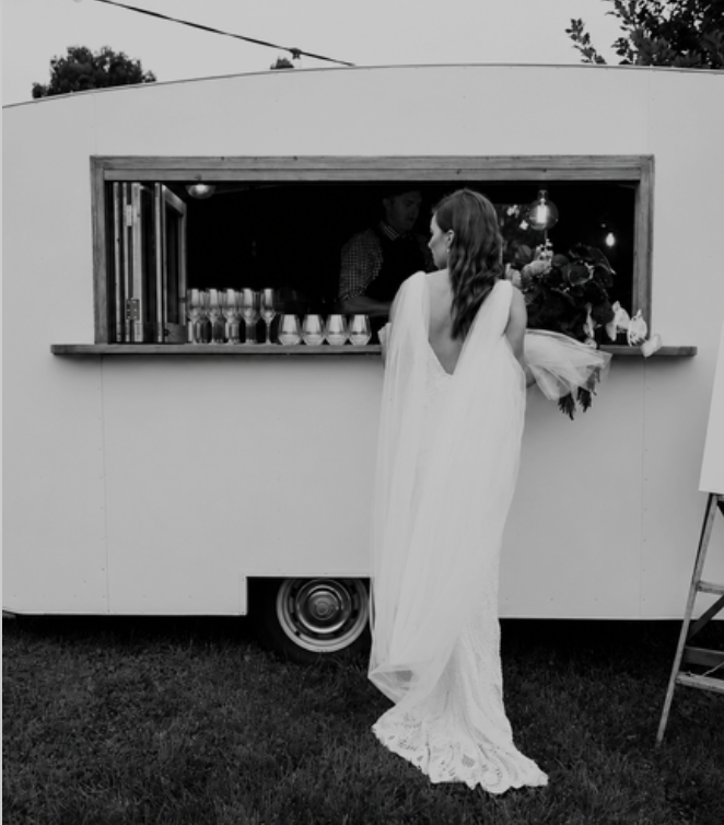 A woman in a wedding dress standing at a food truck or trailer, looking at the person inside who is serving behind the window. There are drinks or containers lined up inside the truck, and a bouquet of flowers is placed on the counter.