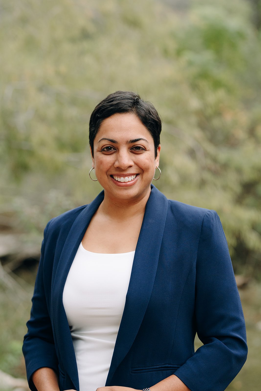 A woman with short black hair, wearing hoop earrings, a white top, and a navy blazer, smiling outdoors with a blurred green background.