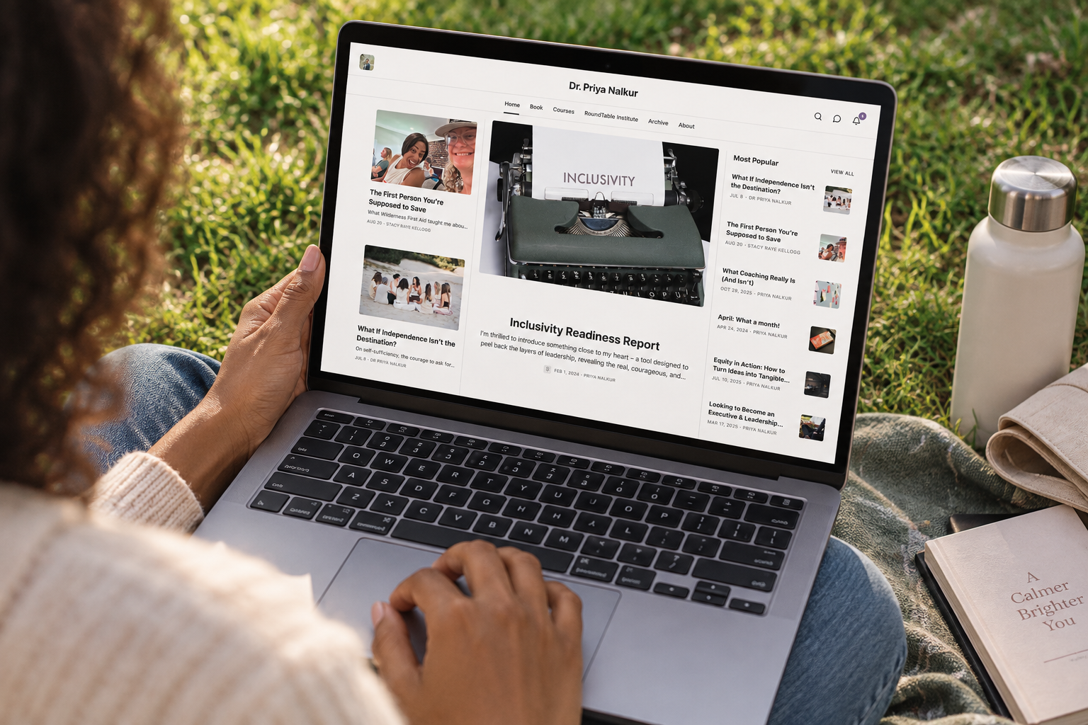 Person holding a laptop open to a webpage titled 'Dr. Priya Nalkur', displaying articles related to inclusivity and leadership, outdoors on a blanket with a notebook and water bottle nearby.