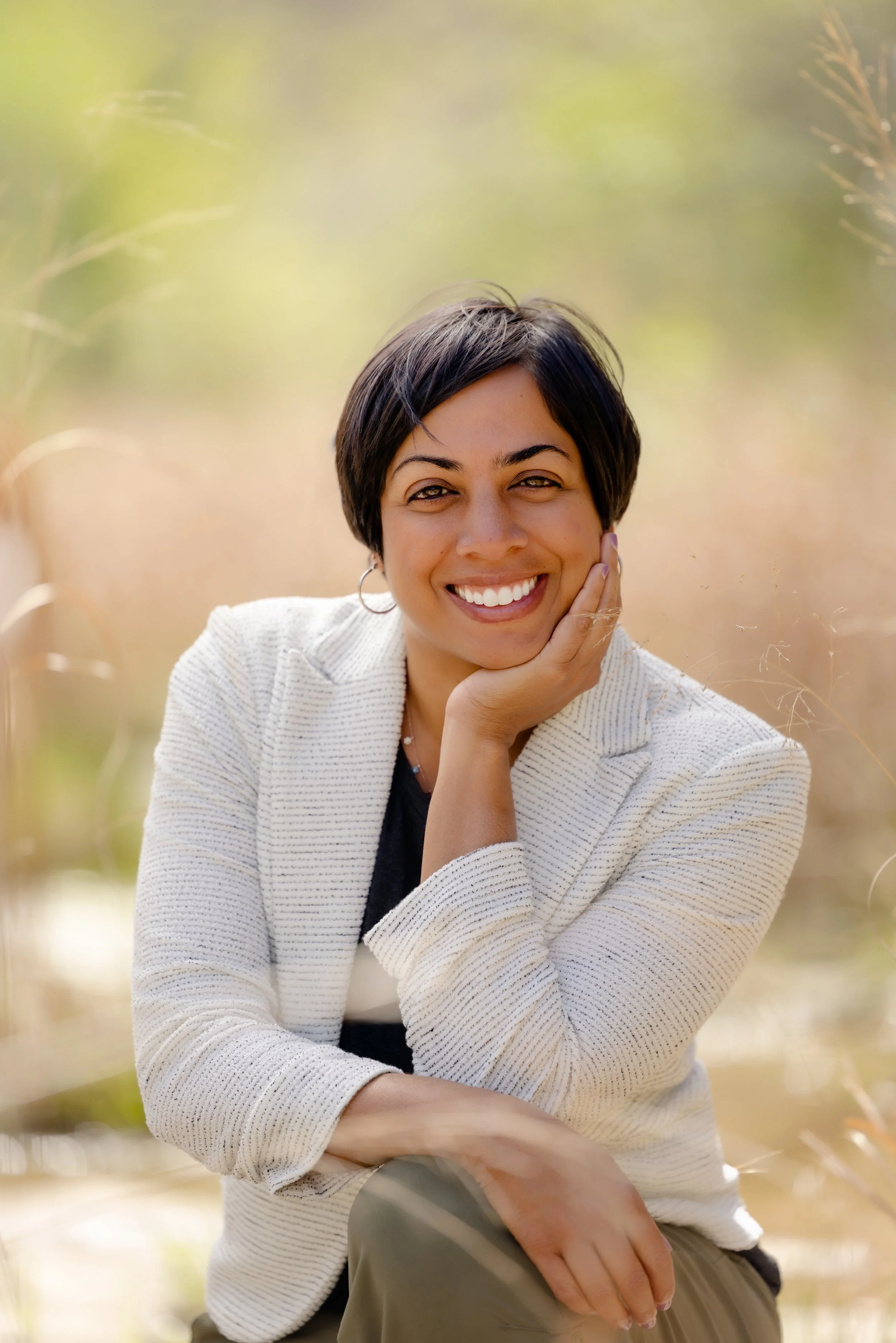 A woman with short dark hair smiling, resting her chin on her hand, sitting outdoors in a natural setting with blurred green and beige background.