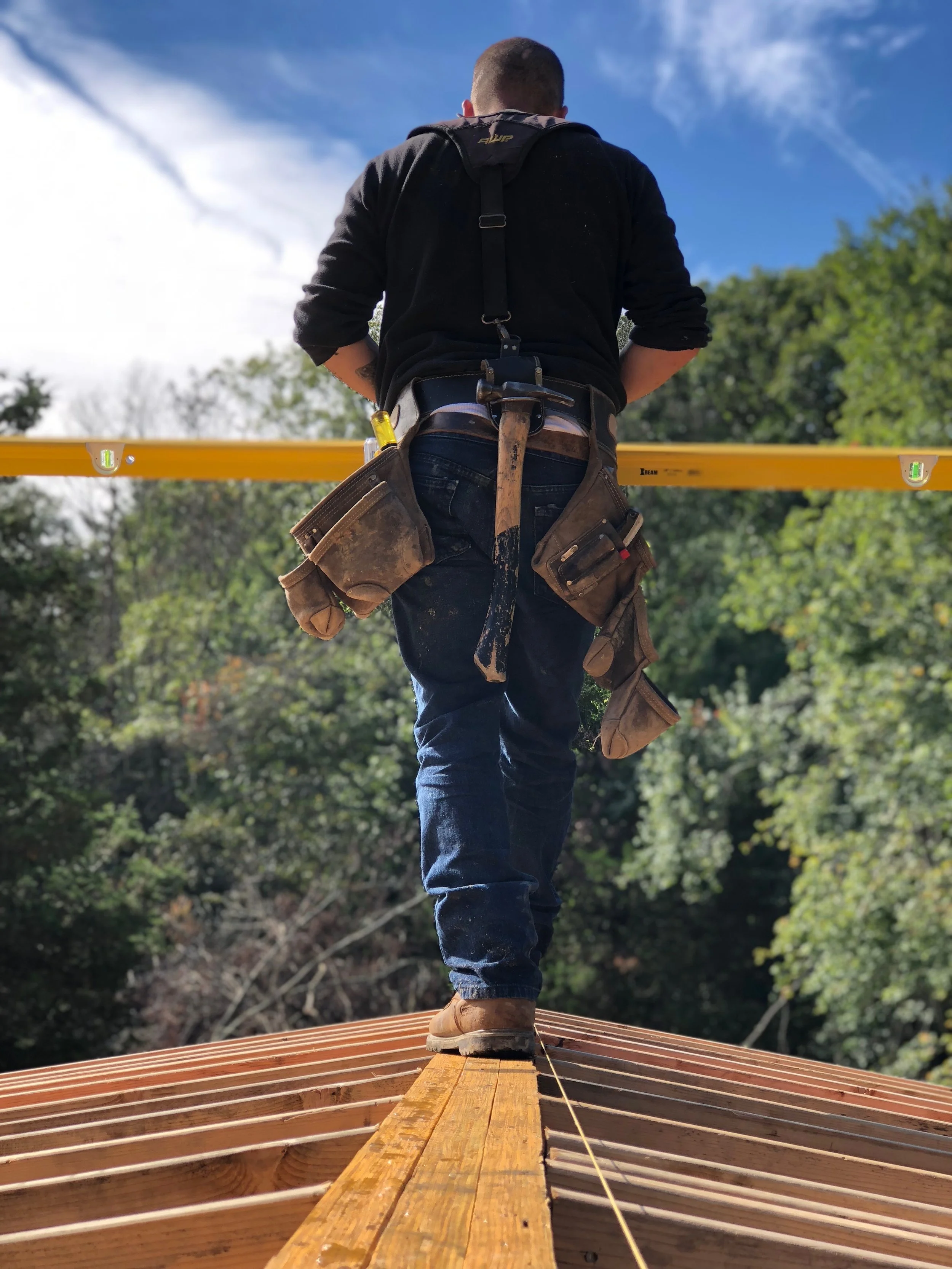 A construction worker standing on a wooden roof structure, facing away, with construction tools attached to his belt, under a blue sky with clouds and trees in the background.